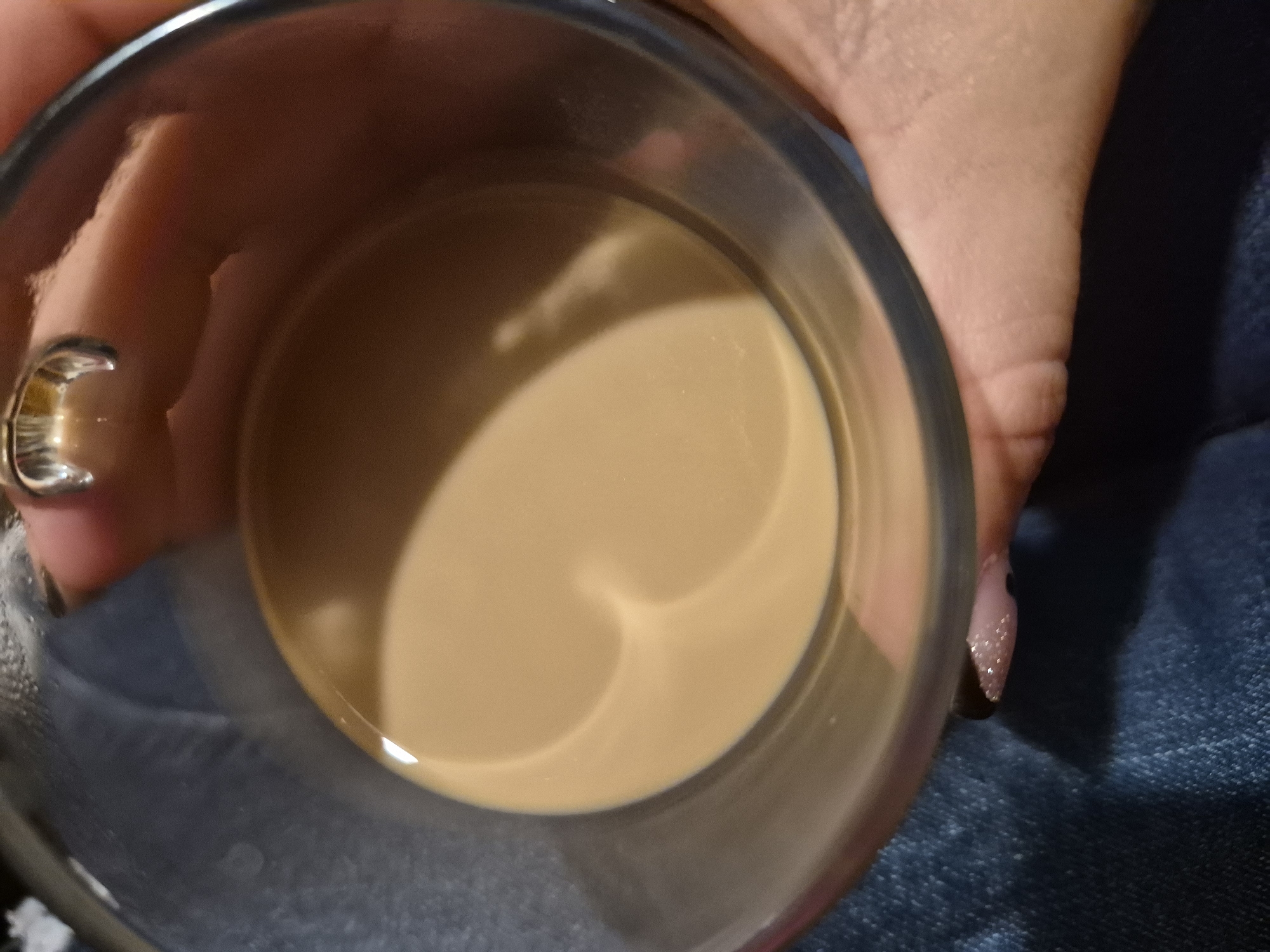 A close-up, overhead shot shows a glass mug filled with a light brown liquid, likely coffee with milk, or tea with milk. The liquid has a swirling pattern on its surface, creating a heart-like shape. A hand, with polished nails, is holding the mug. The background consists of dark blue fabric, possibly jeans. The image does not provide any information or context to determine its location in the city of Sandwell, United Kingdom.