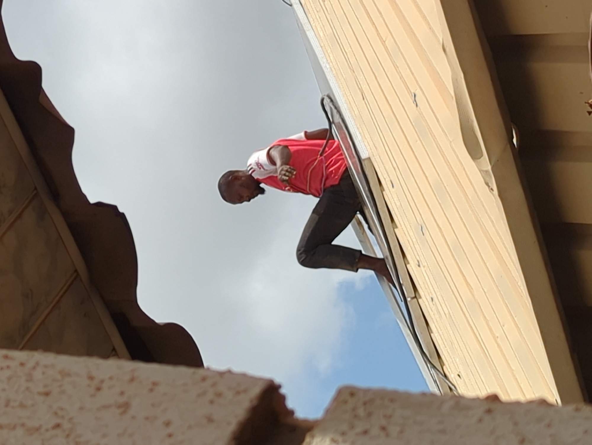 A man in a red shirt and dark pants is climbing a silver ladder leaning against a light brown building. The sky is visible between the building and the ladder, with some clouds. The image is taken from a low angle, looking up, with parts of another building's structure in the foreground. The location is Sabon Tasha, Nigeria.