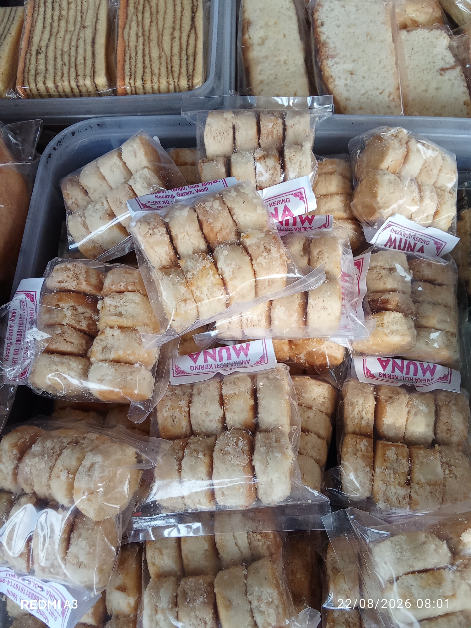 The image shows a close-up of stacked baked goods, likely cookies or small cakes, packaged in clear plastic. These are arranged in several plastic containers, some of which are transparent. The baked goods are light brown and appear to have a slightly crumbly texture, with some pieces dusted with what looks like finely chopped nuts or sugar. Many of the packages have white labels with red text, some of which are partially visible and include "MUNA" and "ANEKA ROTI KERING". There are also some larger slices of cake visible in the background. In the lower right corner of the image, a timestamp "22/08/2026 08:01" is visible. The overall scene suggests a market stall or bakery display. The location is identified as Cemani, Indonesia.
