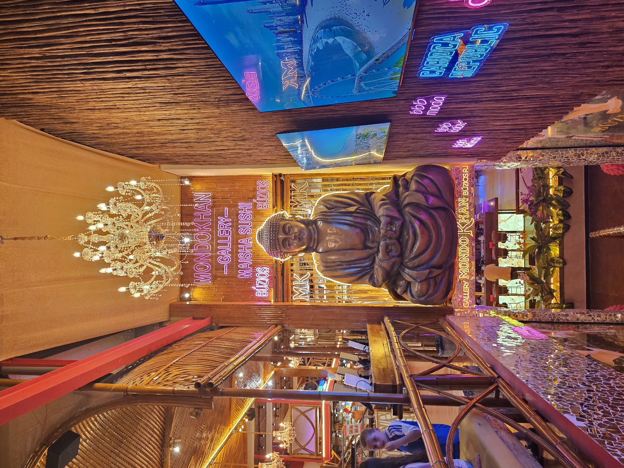 The image depicts the interior of a restaurant or bar located in Armação Dos Búzios, Brazil, based on signage indicating "MONDOKHAN GALLERY-MAISHA SUSHI BÚZIOS." A large Buddha statue dominates the scene. Neon signs are present, including "CARIOCA," "MODA," and other illegible text. The ceiling is made of a textured, wooden material. A chandelier hangs on the left side of the image. A screen displays images of a city, beach, and shark. The decor is elaborate and eclectic.
--- OCR End ---