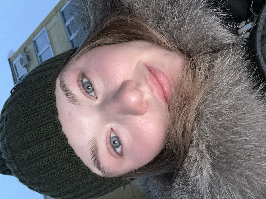 A woman's face dominates the frame, tilted slightly to the side, with light skin, pale pink lips, and blue-gray eyes. She wears a dark green beanie and a fur-trimmed jacket. Part of a light yellow building with snow on the roof and air conditioners is visible in the background, suggesting an outdoor setting. The image is taken at a low angle, looking up at the subject.