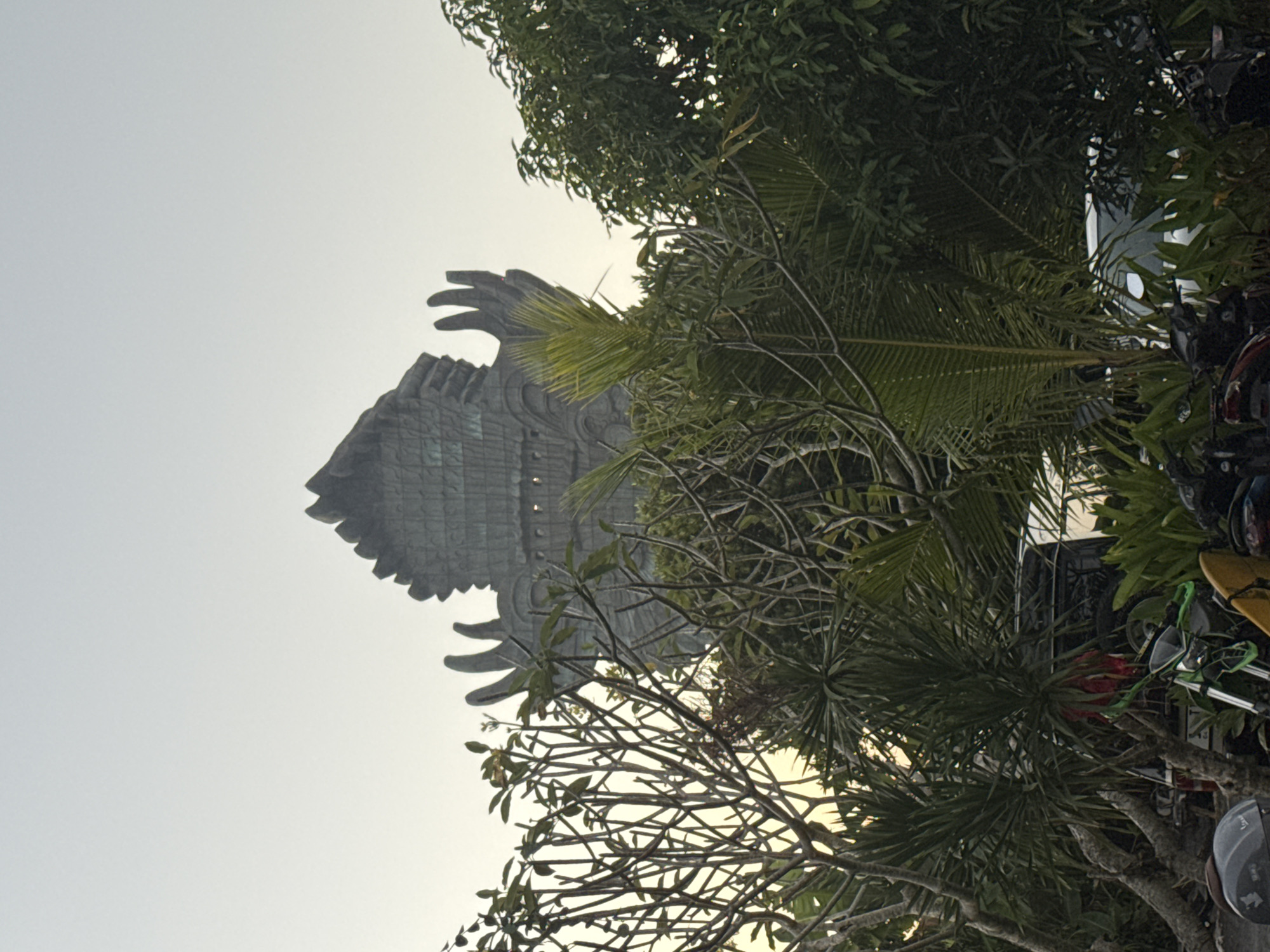 This image shows the Garuda Wisnu Kencana statue in Bali, Indonesia, a prominent landmark in the region of Kuta Selatan. The statue, a colossal structure depicted as a mythical bird-like creature with outstretched wings and a headdress, is partially obscured by lush green foliage and trees in the foreground. The sky is overcast, suggesting it might be dawn or dusk, with a soft, diffused light. In the lower portion of the image, the tops of several motorcycles and a car are visible, indicating a parking area or a street adjacent to the park. The overall scene depicts a tropical environment with a blend of natural elements and man-made structures.