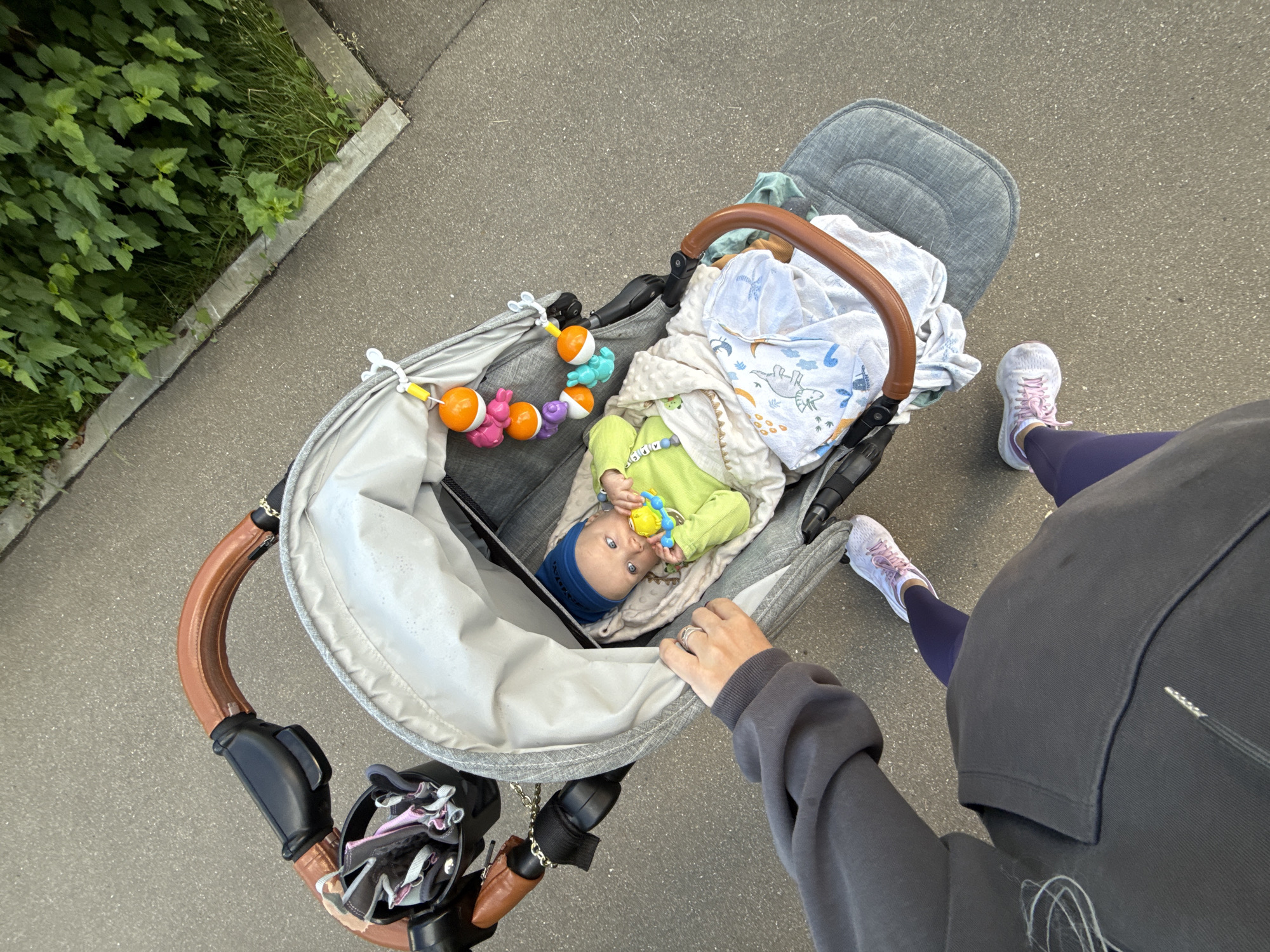 A top-down perspective shot captures a baby in a stroller on a paved surface, possibly a sidewalk or park path. The baby, wearing a blue beanie and a green onesie, lies on its back, chewing on a yellow toy. The stroller is gray and has a brown leather handle. A person's hands are visible, one adjusting the stroller and the other holding a ring on the stroller's frame. The person is wearing a gray sweatshirt and purple leggings, with white sneakers with pink laces. To the right of the paved surface, a strip of green grass and leafy bushes are visible, suggesting an outdoor urban environment. The overall scene depicts a casual outing with a baby. The exact location in Moscow, Russia, is not discernible from the image's visual elements alone.