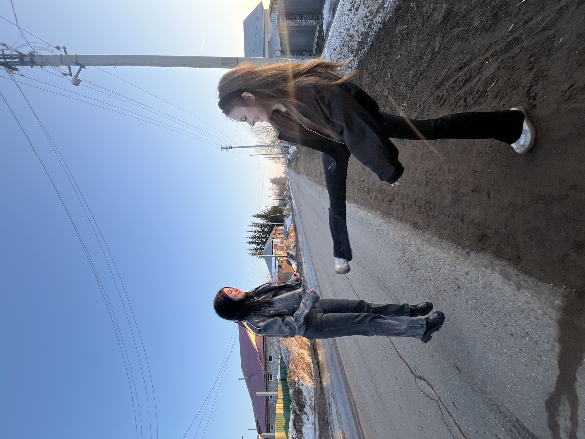 This image depicts two young females engaged in a playful interaction on an asphalt road in the city of Фоки, Russia, under a clear blue sky with low-angle sunlight.
The female on the left, with long dark hair, is seen airborne, approximately 10-15 cm above the road surface, facing towards the right. She wears a dark grey leather-style jacket, dark grey jeans, and black boots, and is smiling with her left arm slightly bent, holding a small dark object.
The female on the right, with long brown hair and a black headband, is positioned on the dirt verge to the right of the road. She is shown executing a high kick with her left leg extended horizontally towards the first female's midsection, while leaning back for balance. She wears a black jacket, black pants, and white and beige sneakers, and is smiling while looking towards the first female.
The asphalt road shows visible cracks and wet patches. Along the left side of the road are piles of dirty snow and mud, fronting a green fence and a stone house with a red-brown roof. On the right side, the muddy dirt verge transitions to a dark metal fence and another house. Multiple utility poles with overhead power lines are visible, extending into the background where bare deciduous trees and evergreen conifers line the street. The environment presents a residential street in a cold or transitional season.