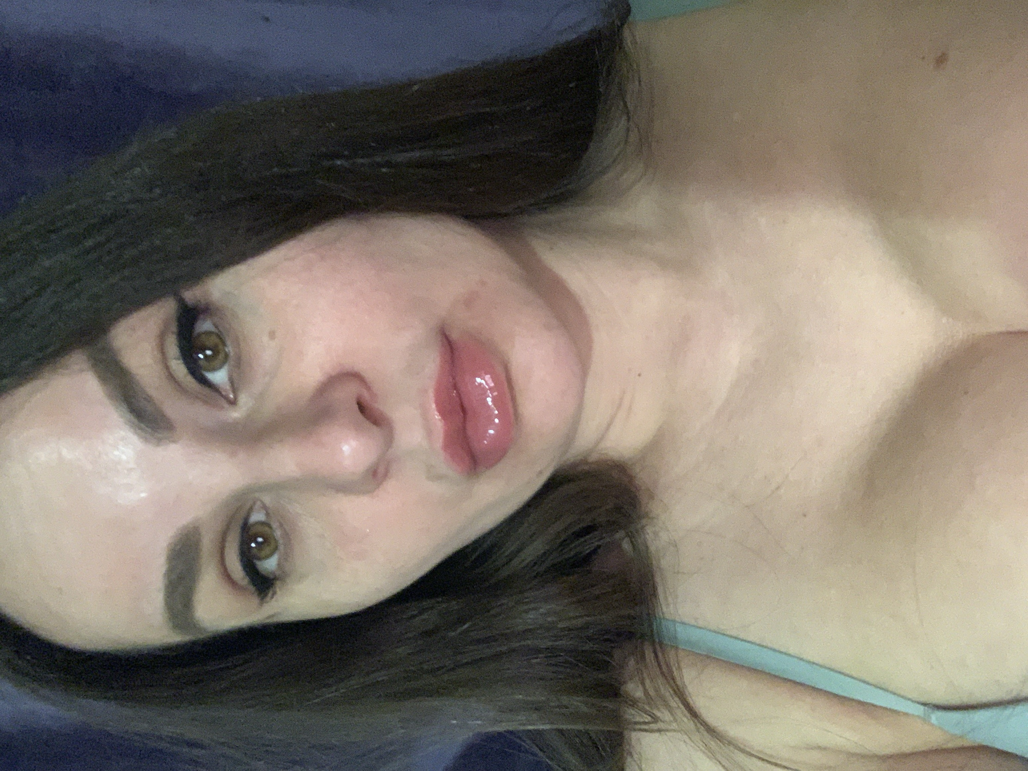 Here's a detailed description of the image:
A close-up portrait shows a woman with long, dark hair and fair skin lying down. Her face is angled towards the camera. She has brown eyes with eyeliner, defined eyebrows, and full lips with a glossy finish. The background consists of blue fabric, likely a bed covering. She is wearing a light blue strap. The location is unknown.