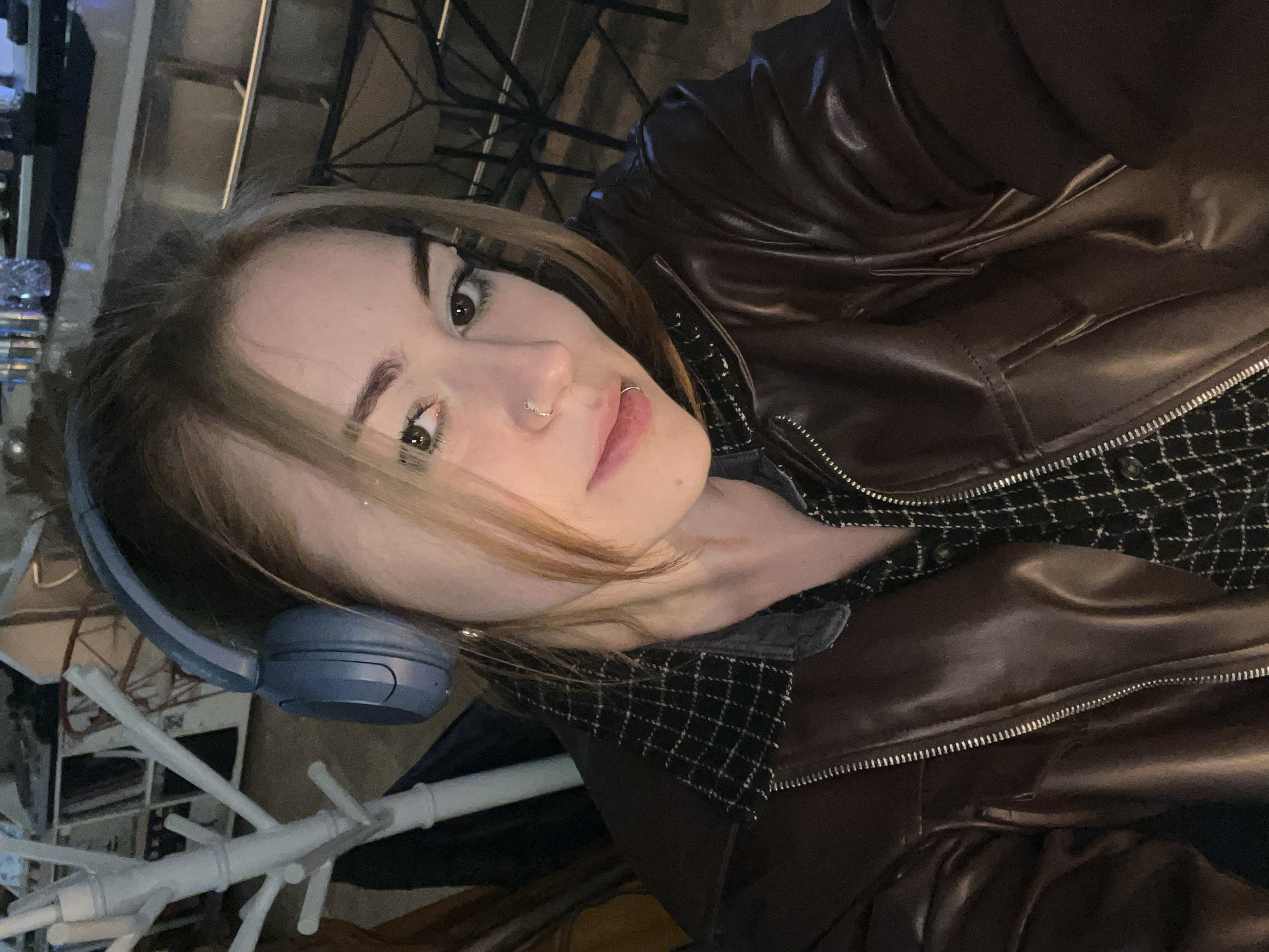 A young adult female is depicted in a medium close-up, facing the camera directly, in an indoor, dimly lit commercial establishment in Rafailovići, Montenegro. She has light brown hair, dark eyes with visible eyeliner, and two silver facial piercings: a hoop in her left nostril and a labret stud on the lower lip. She wears blue over-ear headphones. Her attire consists of a dark brown leather or faux leather jacket with a partially unzipped silver zipper, worn over a black and white plaid shirt with a dark, possibly denim, collar. To her left, a white, multi-pronged coat rack is visible. The right side of the background features a dark counter or bar surface with blurred glass objects and indistinct dark metal furniture below. No discernible actions or interactions are observed.