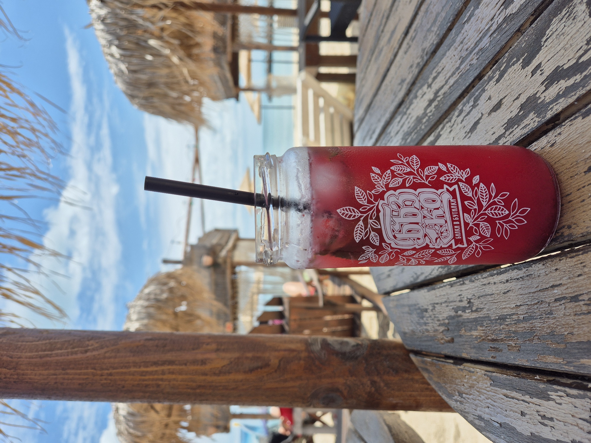A close-up, eye-level shot shows a tall, clear glass jar filled with a red beverage and ice. A dark straw extends from the top of the drink. The jar has a white decorative label featuring Cyrillic text and floral designs. The jar sits on a weathered, grey wooden table with visible grain and peeling paint. In the background, a beach scene unfolds under a bright blue sky with scattered clouds. Several thatched umbrellas and beach chairs are visible, as is the ocean in the distance. A thick, brown wooden pillar is positioned to the left of the jar, partially obscuring the background. The overall setting appears to be an outdoor beachside establishment.