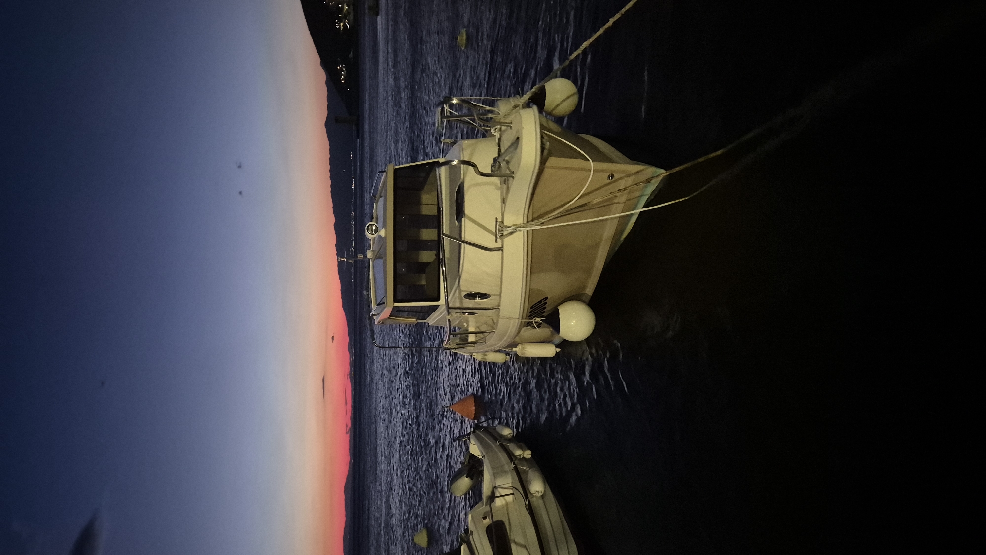 A motorboat is moored at sea during twilight. The boat is light-colored with a closed cabin and metal railings. It is tied to a dock with ropes and has several white fenders attached to its sides. Another smaller boat is moored to the left of the main boat. The water is dark and choppy, reflecting the colors of the sky. The sky transitions from deep blue at the top to a vibrant pink and orange band near the horizon, indicating sunset or sunrise. Silhouetted mountains and buildings are visible in the distance along the coastline. A lighthouse or tower is present on the distant shore. The scene is dark, with some artificial lighting on the boats and shore.