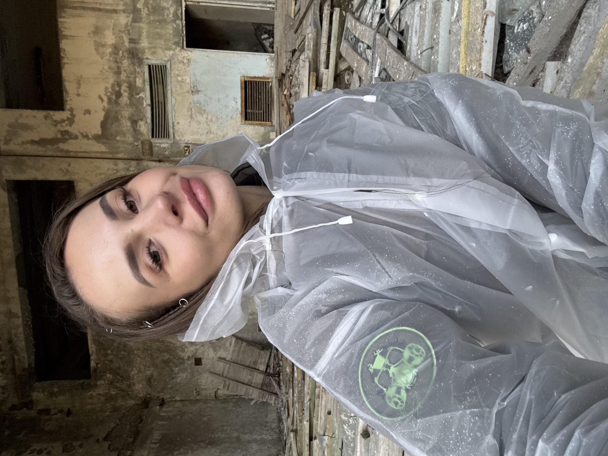 A woman stands inside an abandoned building in Prypiat, Ukraine. The building's interior shows signs of disrepair and decay, with crumbling walls and floors. The woman wears a transparent raincoat with a logo of a radioactive symbol on the arm. The floor is covered with rubble and debris.