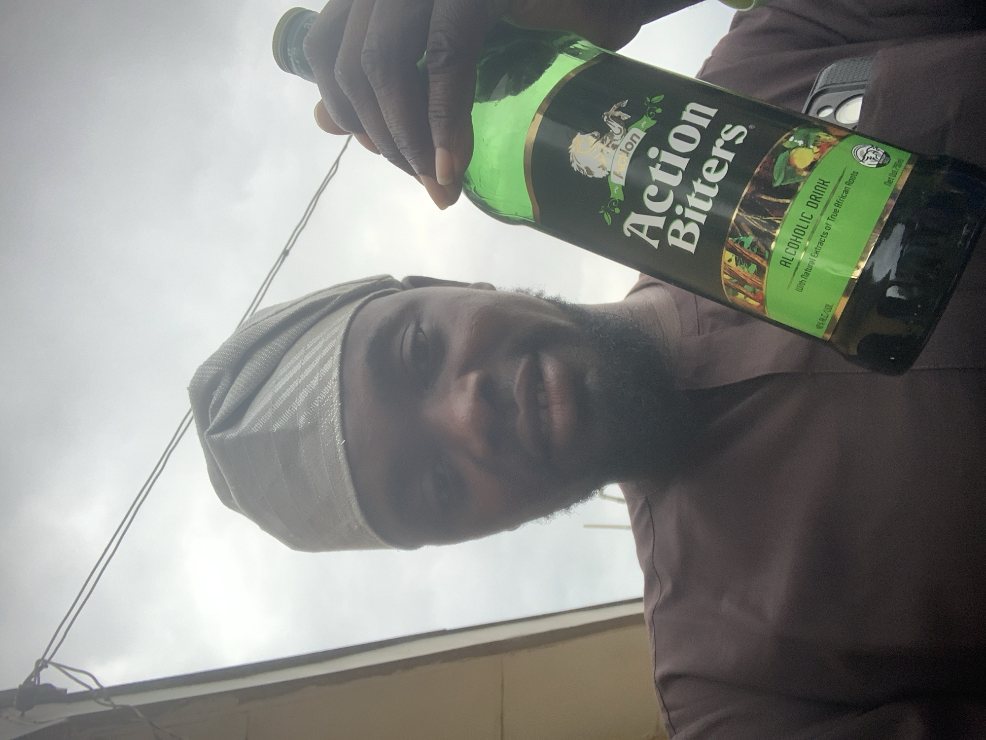 A man, wearing a brown traditional outfit and a light-colored cap, holds up a green bottle of "Action Bitters" alcoholic drink. The bottle label is visible, showing the brand name, "ALCOHOLIC DRINK," and text stating "With Natural Extracts of True African Roots." The man is looking at the camera, and his face is partially illuminated by natural light. He has a short beard. The background is an overcast sky, with a hint of a structure on the left, possibly a building with an overhang. The image appears to have been taken outdoors, suggesting a setting in a town or city, with the brand of bitters suggesting a location in Nigeria, potentially Shagamu.