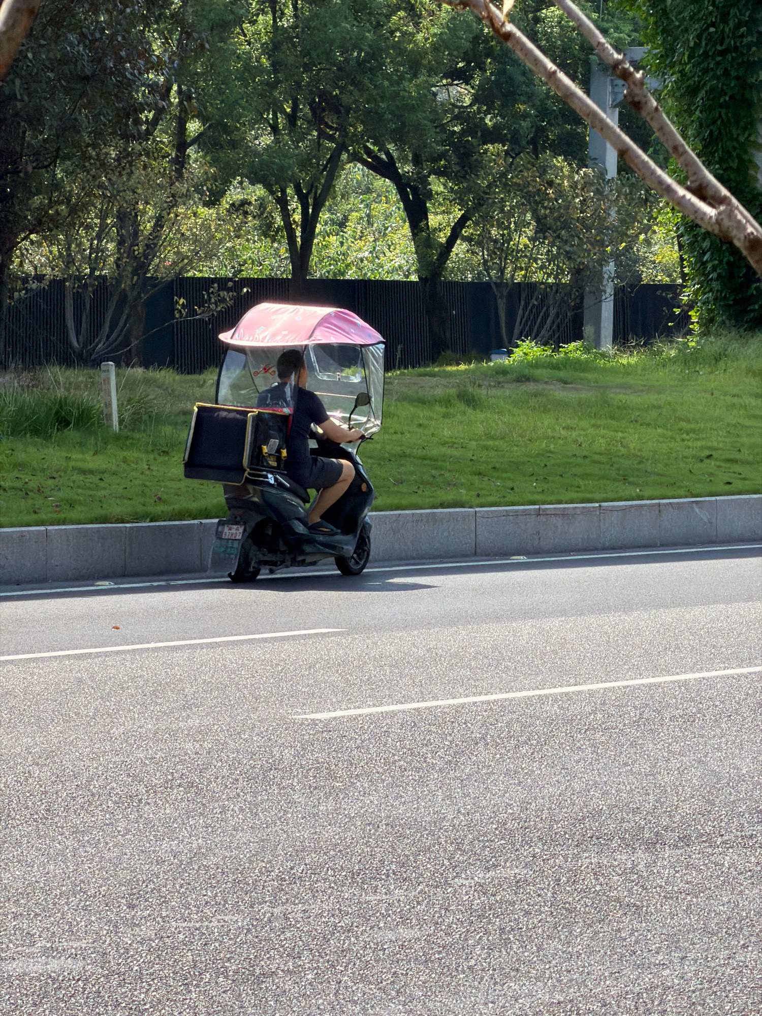 In the city of Jiangjin, China, on a sunny day, a person is riding a dark-colored electric scooter. The scooter has a clear windshield and a pink canopy overhead, suggesting protection from the sun or rain. Attached to the rear of the scooter is a large black cargo box, likely for deliveries. The rider is wearing a dark t-shirt and shorts. The scooter is on an asphalt road, with a gray concrete barrier separating it from a grassy embankment. Behind the embankment, a dark fence and lush green trees are visible, indicating an outdoor urban or suburban setting. The scene appears to be captured from a slightly elevated perspective, looking down at the scooter and the road.