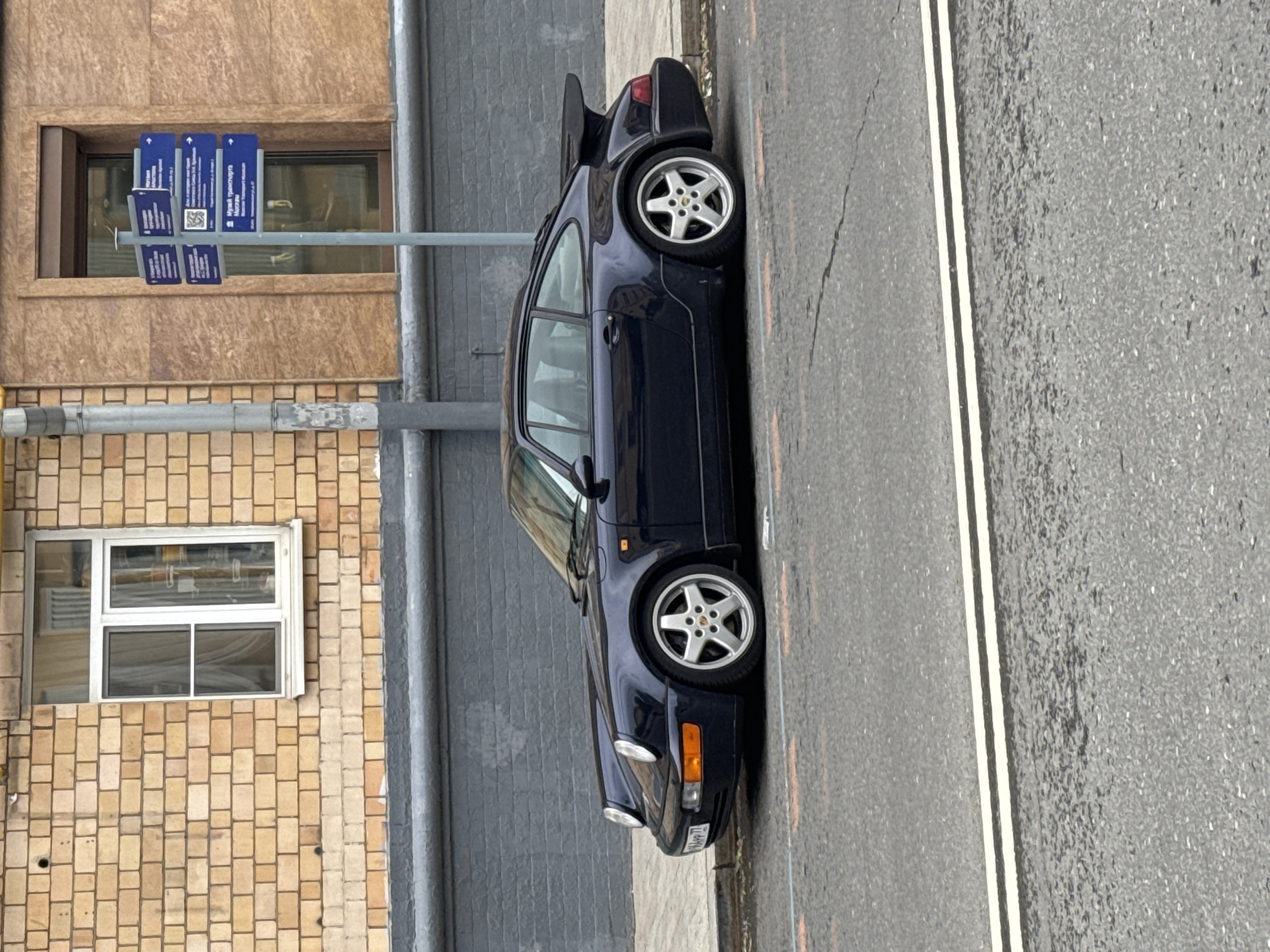 The image shows a dark blue Porsche 911 parked on a city street. The car is facing forward, and its wheels are visible. In the background, there is a brick building with windows and signs. The location is Moscow, Russia, as indicated by the signs which translate to "Moscow Transport Museum". The road markings include double solid lines. No people are visible in the frame.