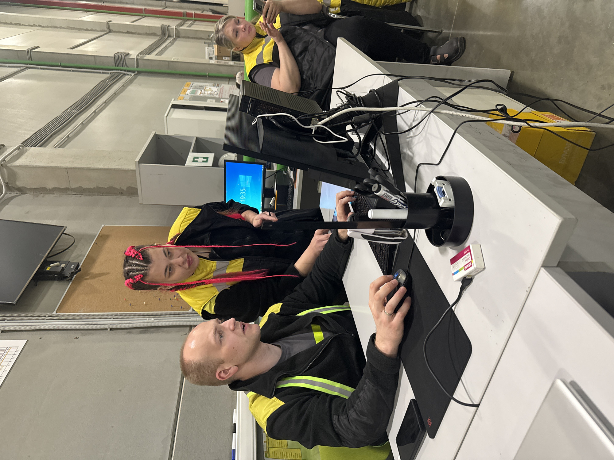 The image depicts three individuals in what appears to be an industrial or warehouse setting in Lupanovo, Russia. Two individuals, a man and a woman, are positioned at a white countertop, likely a workstation. The man is seated and operating a computer mouse on a black mousepad. He is wearing a black jacket with neon yellow stripes. The woman is standing beside him, partially visible, and is wearing a similar jacket with neon yellow stripes and has pink braids. A desktop computer monitor, keyboard, and other office supplies are present on the workstation. A third individual, a woman, is seated behind the workstation, holding a mobile phone. This woman is also wearing a similar jacket with neon yellow stripes.
The background includes concrete walls with exposed conduits, a gray cabinet with a green cross symbol, and a corkboard. The overall environment suggests an office area within a larger industrial space. The presence of the individuals in similar jackets indicates they may be coworkers. The orientation of the image is rotated counterclockwise.