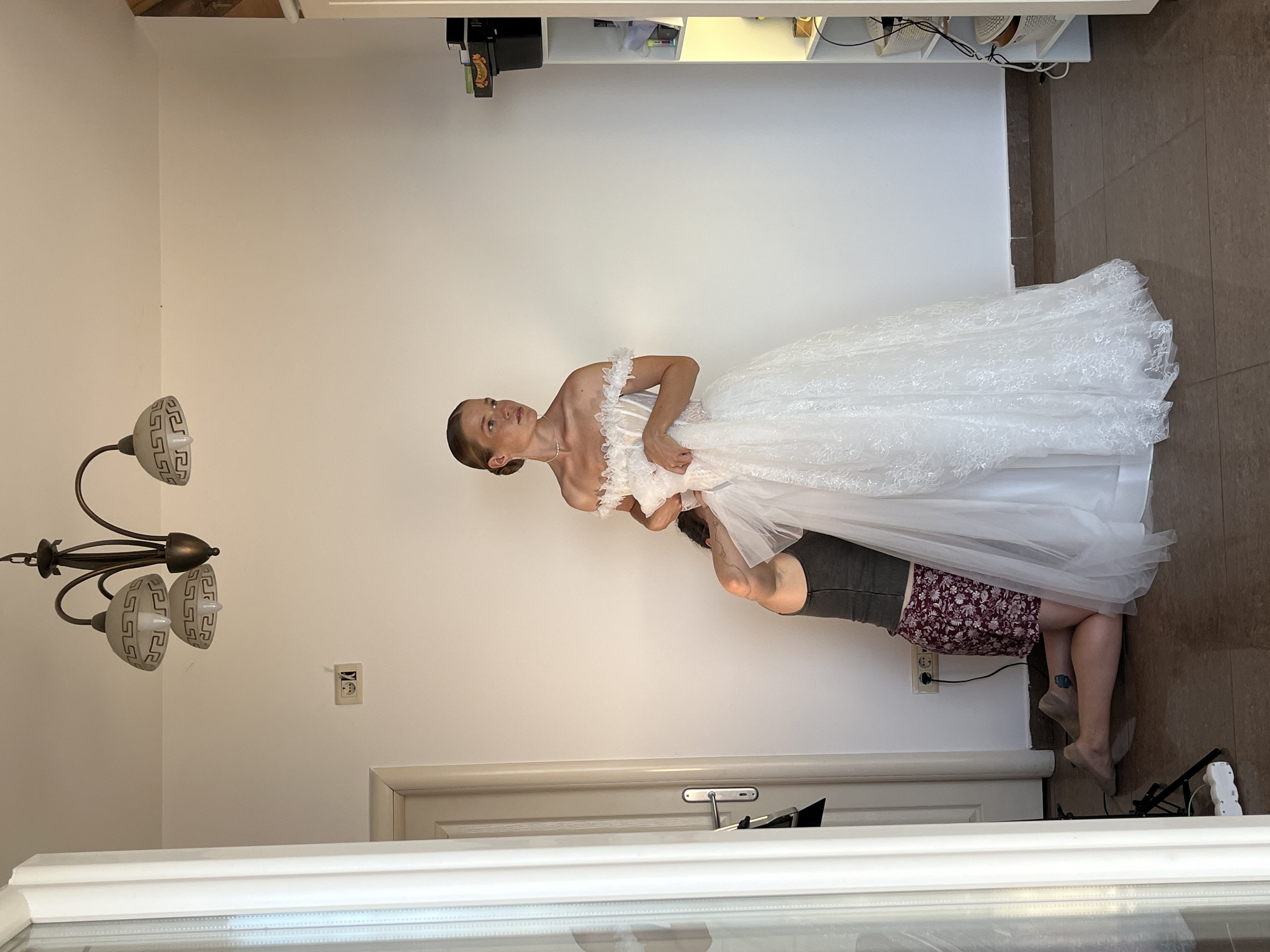 In an indoor setting, possibly a room in Budva, Montenegro, a young woman stands wearing a white, off-the-shoulder wedding dress with lace detailing. She is looking to her right. To her left, a person is kneeling and appears to be adjusting the dress. The kneeling person wears a dark tank top and a patterned skirt. The environment includes a white wall, a chandelier on the ceiling, a door with a handle, electrical outlets, and a white shelving unit with baskets on the right side. The floor is tiled.