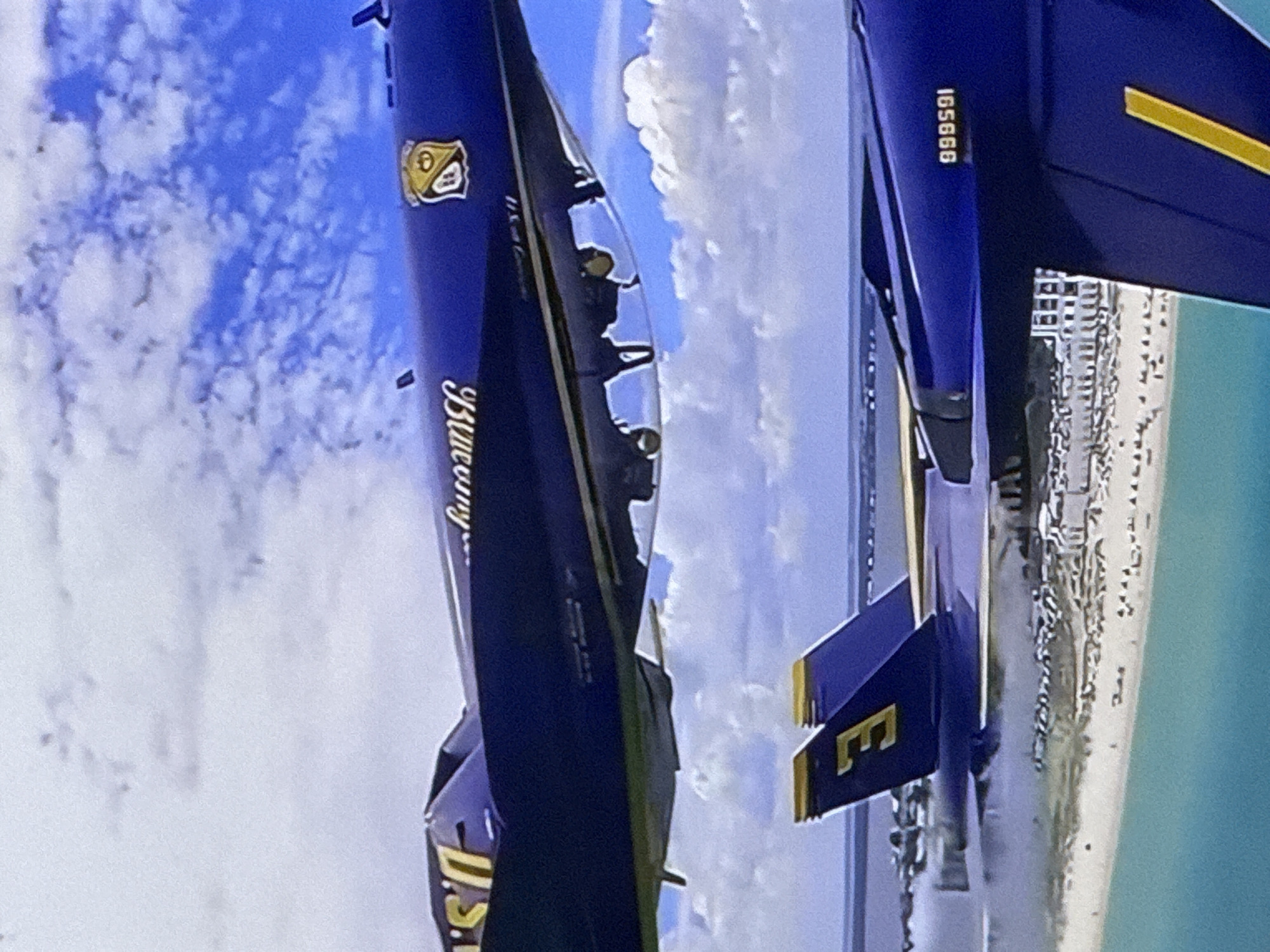 The image shows two blue fighter jets flying in formation against a bright blue sky with scattered white clouds. The jets have yellow accents on their wings and tails. One jet is closer to the viewer and displays the word "Bluey" written vertically in white lettering, along with "U.S. NAVY" on the bottom. The other jet, further back, has the number "165668" printed vertically on its fuselage. Both jets have pilots visible in their cockpits.
In the background, a coastal city with numerous buildings is visible along a sandy beach, bordering a turquoise body of water. The overall scene suggests an airshow or a training exercise taking place on a clear, sunny day. The orientation of the image is vertical, making it appear as if the jets are flying upside down or at a steep angle relative to the viewer. No specific time of day or weather conditions beyond "clear and sunny" can be inferred. The location context provided, Cahul, Moldova, is not visually supported by any elements within the image itself, which instead suggests a coastal setting.