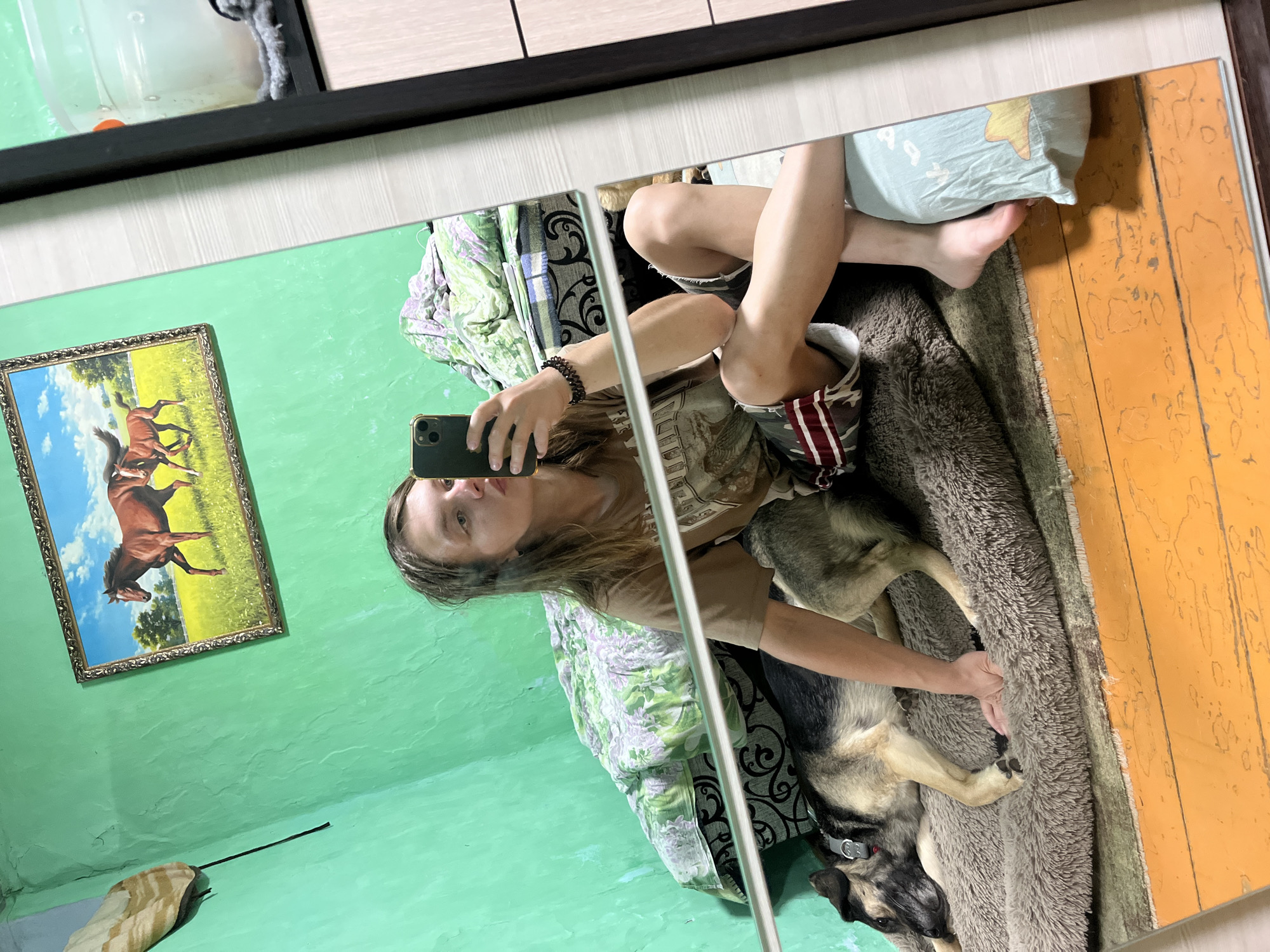 A young woman with long brown hair takes a mirror selfie in what appears to be a bedroom. She is wearing a brown t-shirt and patterned shorts, with a beaded bracelet on her left wrist. She holds a smartphone in her right hand, angled to capture her reflection. To her left, a German Shepherd-mix dog lies on a fluffy brown dog bed, with its head resting on the edge. The room has green walls, and a framed picture of horses hangs on one wall. A bed with green and white floral bedding is visible in the background. The floor is made of orange-painted wood. A portion of a closet or cabinet is visible on the right side of the frame. The image quality suggests it was taken indoors with artificial lighting. The scene is domestic and casual.