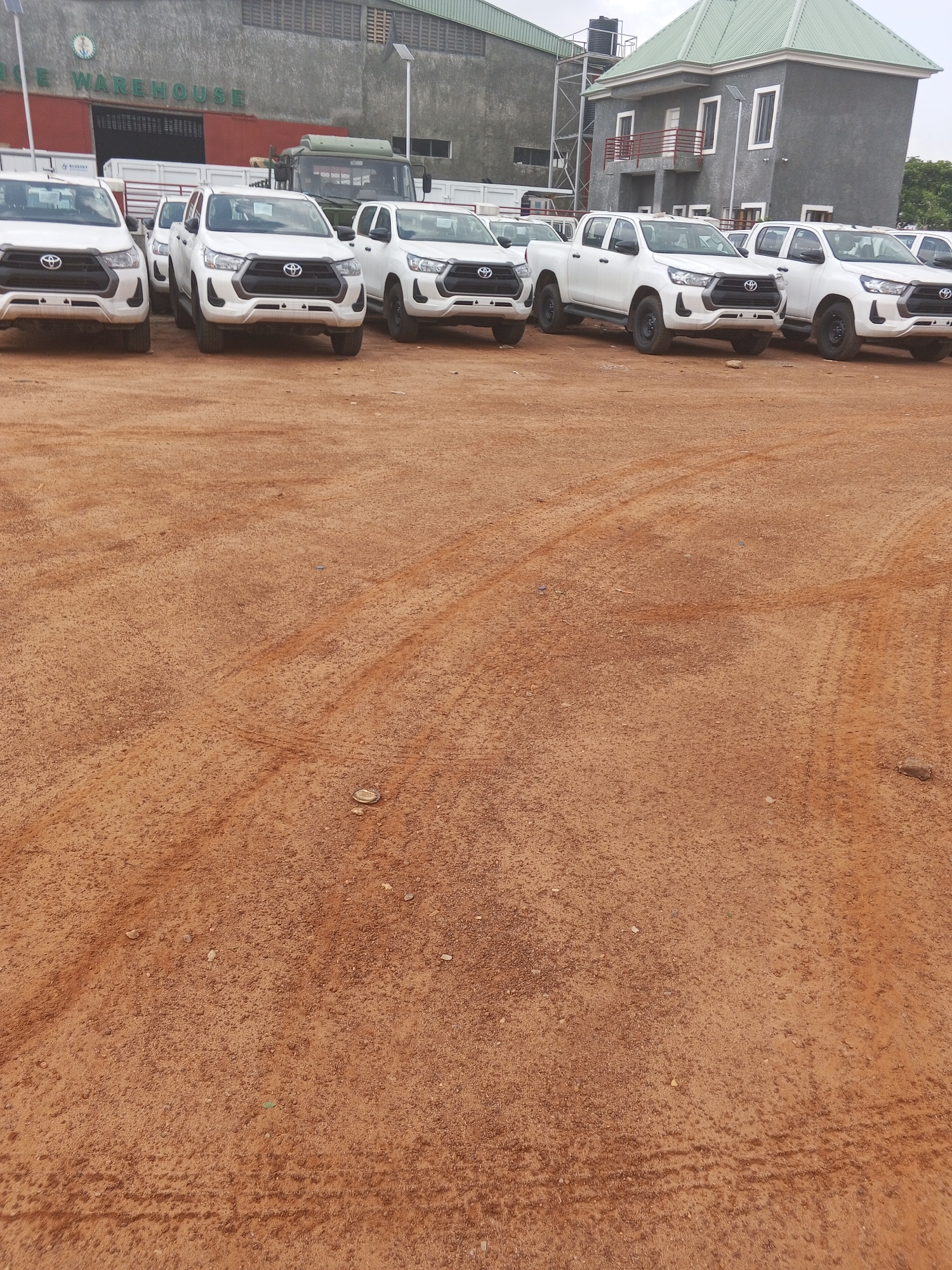 The image depicts a row of white Toyota Hilux pickup trucks parked on a reddish-brown, dirt surface in what appears to be a staging area or lot. The location is indicated by the presence of a building with signage that reads "Rice Warehouse," suggesting an industrial or commercial district. In the background is another grey-colored building with a greenish-grey roof. There is a large truck parked behind the Hilux vehicles, and the ground shows tire tracks, indicating vehicle movement. The sky is overcast. The location is likely Abuja, Nigeria, due to the architectural style of the buildings and the coloring of the soil, which are characteristic of the region.