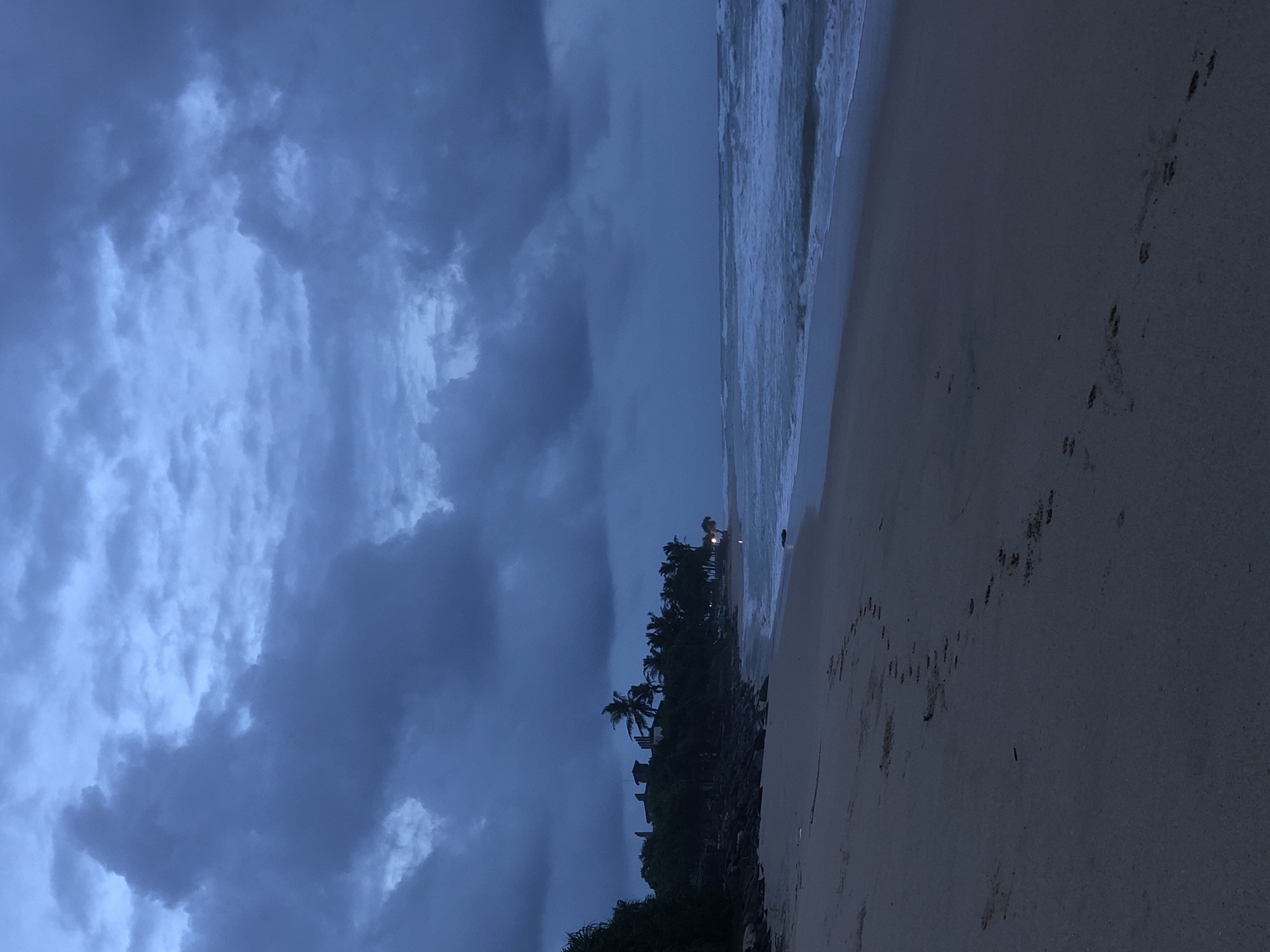 The image depicts a beach scene at dusk in Kathaluwa, Sri Lanka. The sky is filled with dark, heavy clouds, suggesting an impending storm or recent rainfall. The sea is moderately rough, with waves breaking onto the sandy shore. In the distance, along the coastline, palm trees and some buildings are visible, silhouetted against the dim light. Footprints are scattered across the wet sand in the foreground, indicating recent human presence. The overall atmosphere is somber and moody, characterized by the twilight lighting and the dramatic cloud formations.