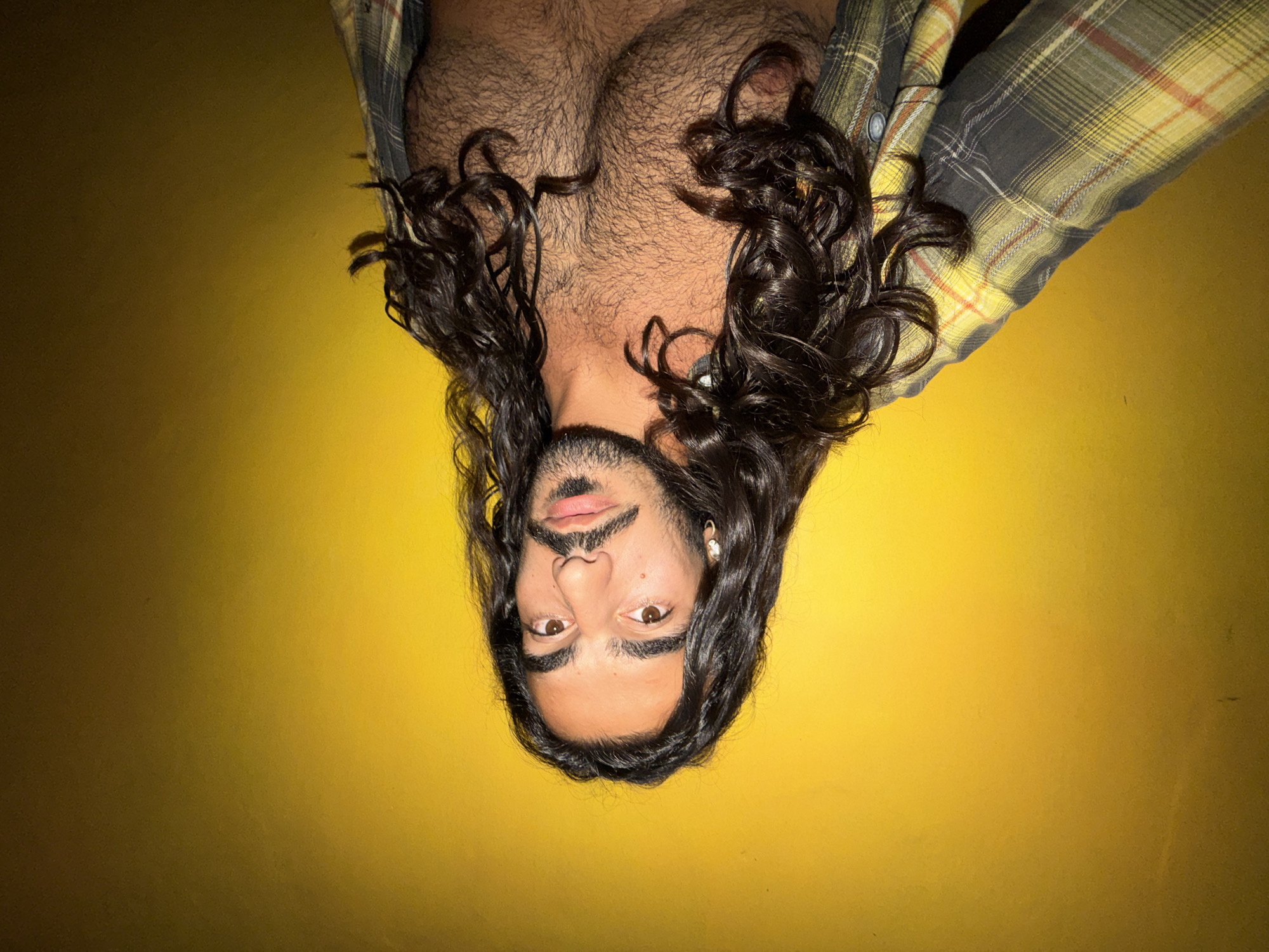 The image is an angled, close-up shot of a man with long, dark, curly hair and a beard, wearing a plaid shirt. The background is a solid yellow wall. The man's face is turned towards the camera, and he has light brown eyes and dark eyebrows. He is wearing a silver hoop earring in his left ear. His chest is visible and appears to have dark hair. The lighting is somewhat dim, casting shadows on the wall and highlighting the texture of the man's hair and skin. The photo appears to be taken indoors. There is no indication of location in North Kuta, Indonesia within the image itself.