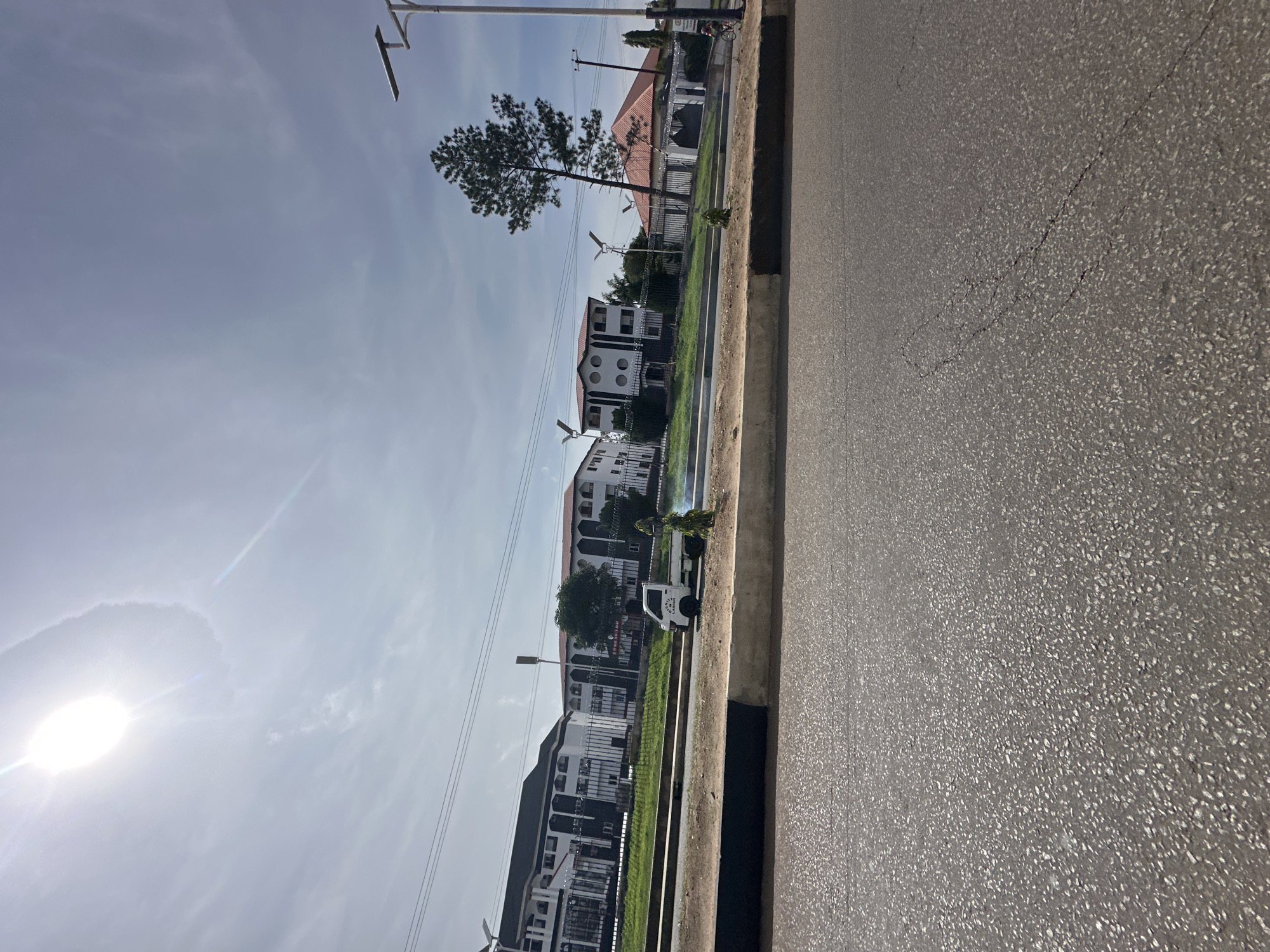 The image shows a sunlit street scene in Bukuru, Nigeria. In the foreground, a paved road with visible cracks is visible. Beyond the road, a raised median with a black and white curb separates it from a grassy area. On this grassy area, a small white truck with "UNITY CARGO" printed on its side is parked. To the right of the truck, a small evergreen tree is visible, with some green smoke or dust emanating from its base.
Behind the median, a row of modern buildings with white and dark facades are situated behind a metal fence. One building has a distinctive red-tiled roof. A tall evergreen tree stands in front of one of the buildings. In the sky, the sun is brightly shining, creating a lens flare effect and casting a halo. Power lines stretch across the upper part of the image. A street lamp is visible on the right side of the frame. A person on a bicycle is visible in the far right background, riding along the side of the road.
