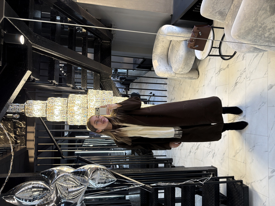 An indoor mirror selfie taken by a female individual in a modern, upscale commercial interior in Moscow, Russia. The woman, with light brown hair, is wearing a long dark brown coat, a cream-colored fringed scarf, and black pointed-toe boots. She holds a silver smartphone in her right hand, angled towards her reflection.
The environment features a white marble-patterned tiled floor and a black ceiling. A large, illuminated multi-tiered crystal chandelier hangs prominently. To the left, a black metal open-tread staircase with railings is partially obscured by a cluster of silver metallic foil balloons.
Reflected in the mirror on the right is a light grey textured armchair positioned next to a small round table with a marble top. A brown leather handbag with a silver clasp rests on the table. Further reflections show an office chair and dark vertical room dividers, indicating an adjacent space. The setting conveys a contemporary, luxurious aesthetic.