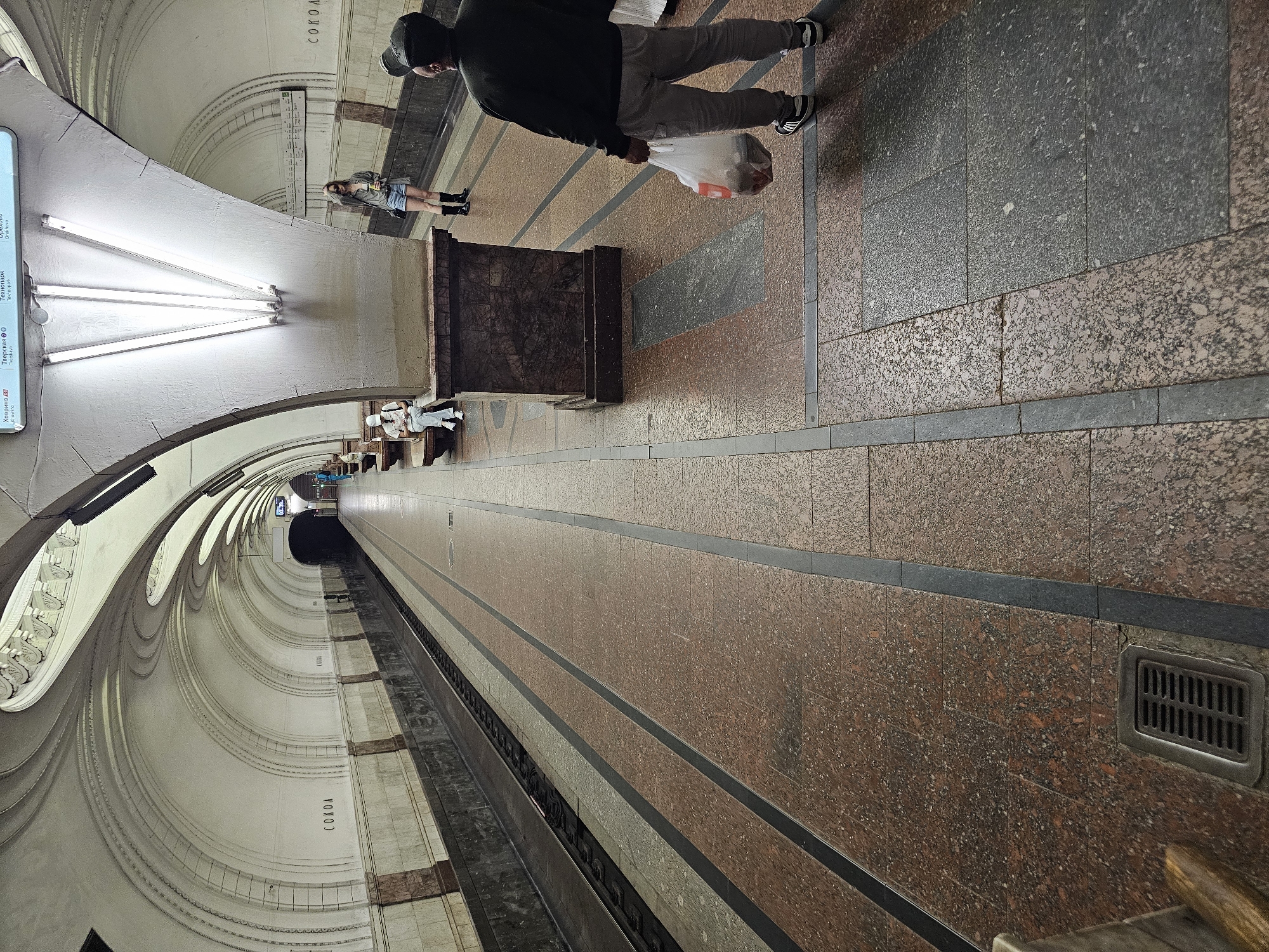 The image depicts a subway platform in the city of Khimki, Russia. The platform is constructed with granite tiles in varying shades of red and gray, with black granite accents and a metal grate drain in the foreground. The architecture features high, arched ceilings with decorative molding and long rows of fluorescent lights. The walls are lined with marble and have the word "СОКОЛ" (Sokol) inscribed on them.
There are several individuals on the platform. In the foreground, a person wearing a dark jacket, gray pants, and black and white sneakers is holding a plastic bag filled with items. To the left, a person in a white cap and light-colored clothing sits on a bench. Further down the platform, another person in denim shorts and black boots stands, holding a drink and sunglasses. In the distance, another individual in blue is visible near the tunnel entrance. The platform appears empty and quiet, with a train track running along the left side of the image.