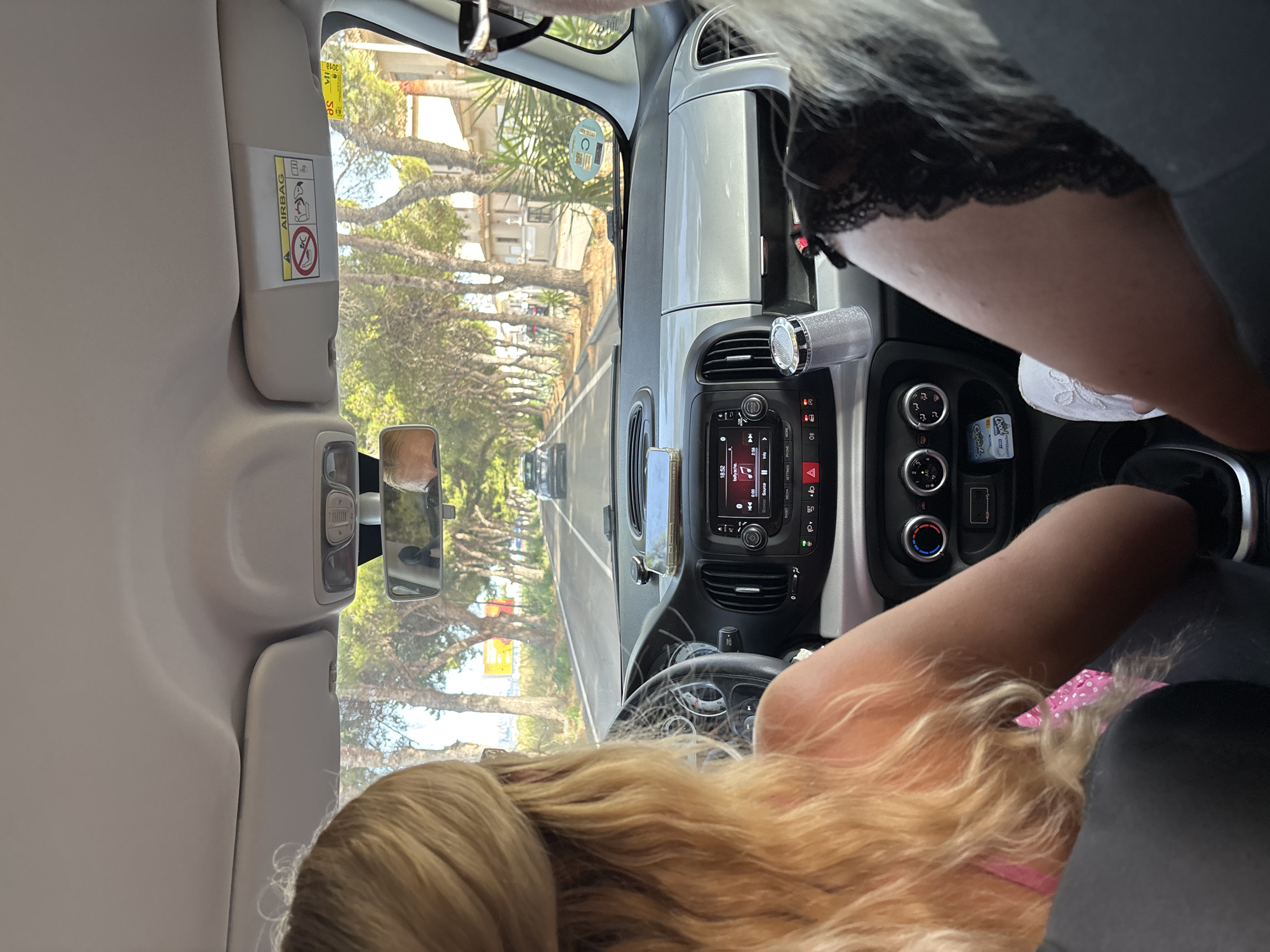 The image shows the interior of a car, viewed from the back seat, looking forward. A blonde woman with her back to the camera is driving, wearing a pink top with white polka dots. Her hair is styled in loose waves. To her right, another person's shoulder and leg are visible, wearing black lace trim and a white shoe. The dashboard features a multimedia screen displaying song information, climate control knobs, and buttons. A silver-colored cup holder with a sparkly container is visible on the dashboard. Through the windshield, a tree-lined road is seen, with a black car ahead. Buildings are visible on the right side of the road. The sky is bright, suggesting daytime. An "AIRBAG" warning sticker is affixed to the car's visor. The overall scene suggests a road trip or drive through a suburban or coastal area, possibly during warm weather.