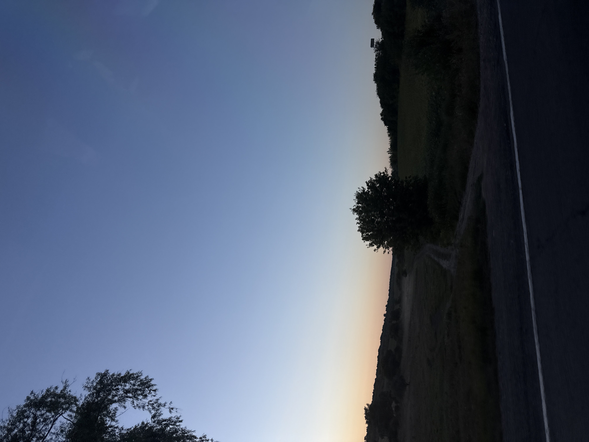 This is a low-angle, full shot of a rural landscape at dusk, likely in the city of Fundul Galbenei, Moldova. The sky transitions from a pale blue at the top to a faint orange near the horizon. In the foreground, a dark asphalt road with a white line marking runs horizontally across the bottom of the frame. To the left of the road, there is a patch of dark grass and foliage. Beyond the road, the land rises into rolling hills covered in vegetation, with a few scattered trees. A dirt path is visible winding its way up one of the hills. In the middle ground, a solitary, dark-leaved tree stands prominently. Further in the distance, more trees and bushes form a dark treeline along the horizon. The overall impression is one of quiet solitude in a natural setting as daylight fades. There are no people, animals, or vehicles visible in the image.