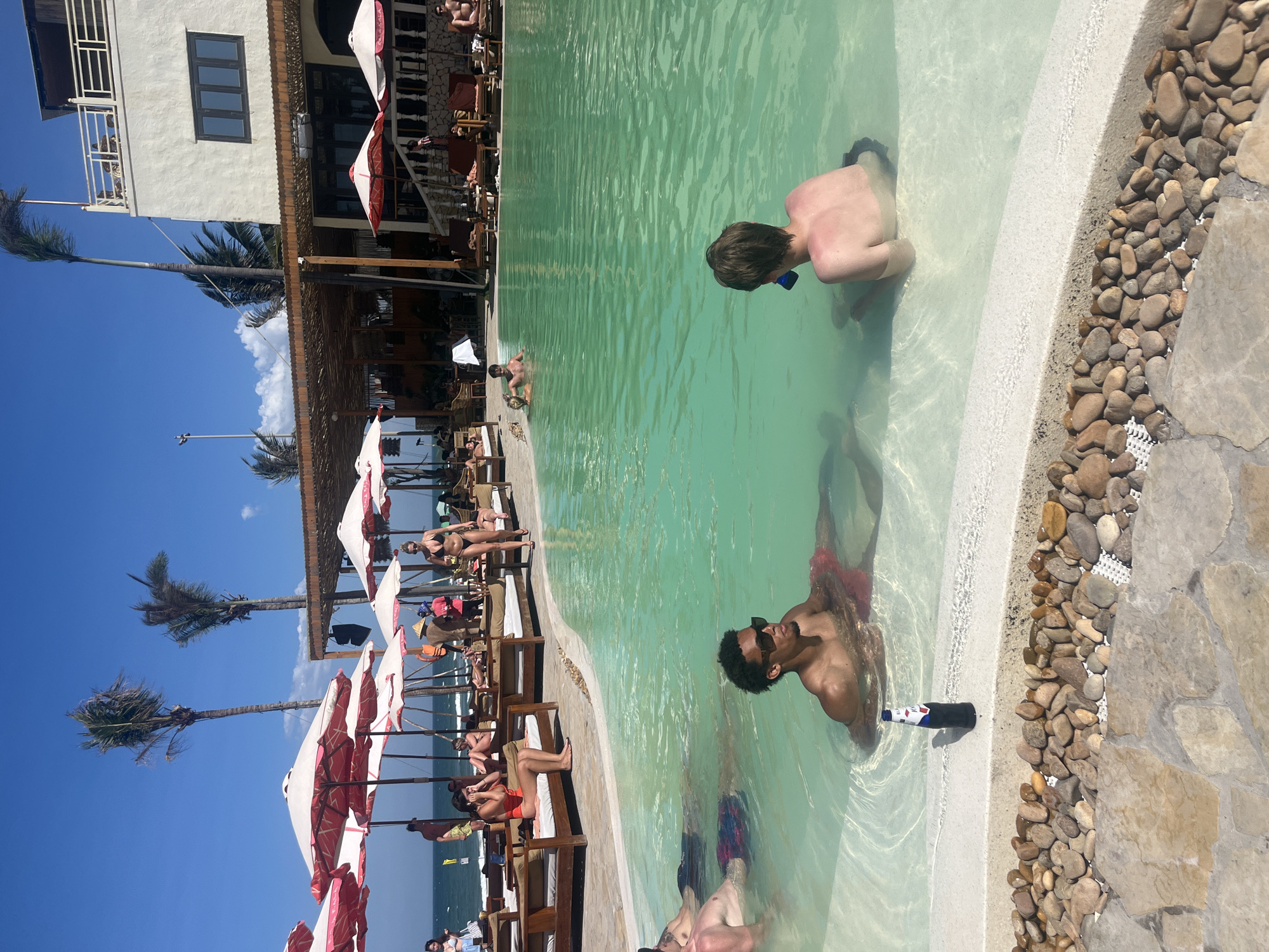 This media file captures a vibrant outdoor scene at a tropical resort or beach club in Đà Nẵng, Vietnam, on a bright, sunny day. The dominant feature is a light green/turquoise swimming pool, where several people are enjoying the water.
In the foreground, two men are prominently featured in the pool. One man, seen from the back, has swim goggles around his neck or face. Another man, with dark hair and sunglasses perched on his head, is partially submerged in the pool, looking towards the left. A dark green bottle, identifiable as a Heineken bottle by its label showing "HEINEKEN" in white text with a red star, floats next to him in the water. Other individuals are also partially visible in the pool.
Around the pool, there's a white concrete edge decorated with natural brown and grey stones, leading to a paved stone area. Beyond the poolside, numerous lounge chairs are arranged under large red and white striped beach umbrellas. Several people are relaxing on these chairs, sunbathing or enjoying the shade.
In the background, a two-story white building with dark windows and a balcony is visible on the left. Tall palm trees punctuate the scene, reaching towards a clear blue sky dotted with a few white clouds. The expanse of the sea can be glimpsed in the distance, adding to the coastal resort ambiance. The overall atmosphere conveys relaxation and leisure in an idyllic sunny setting.