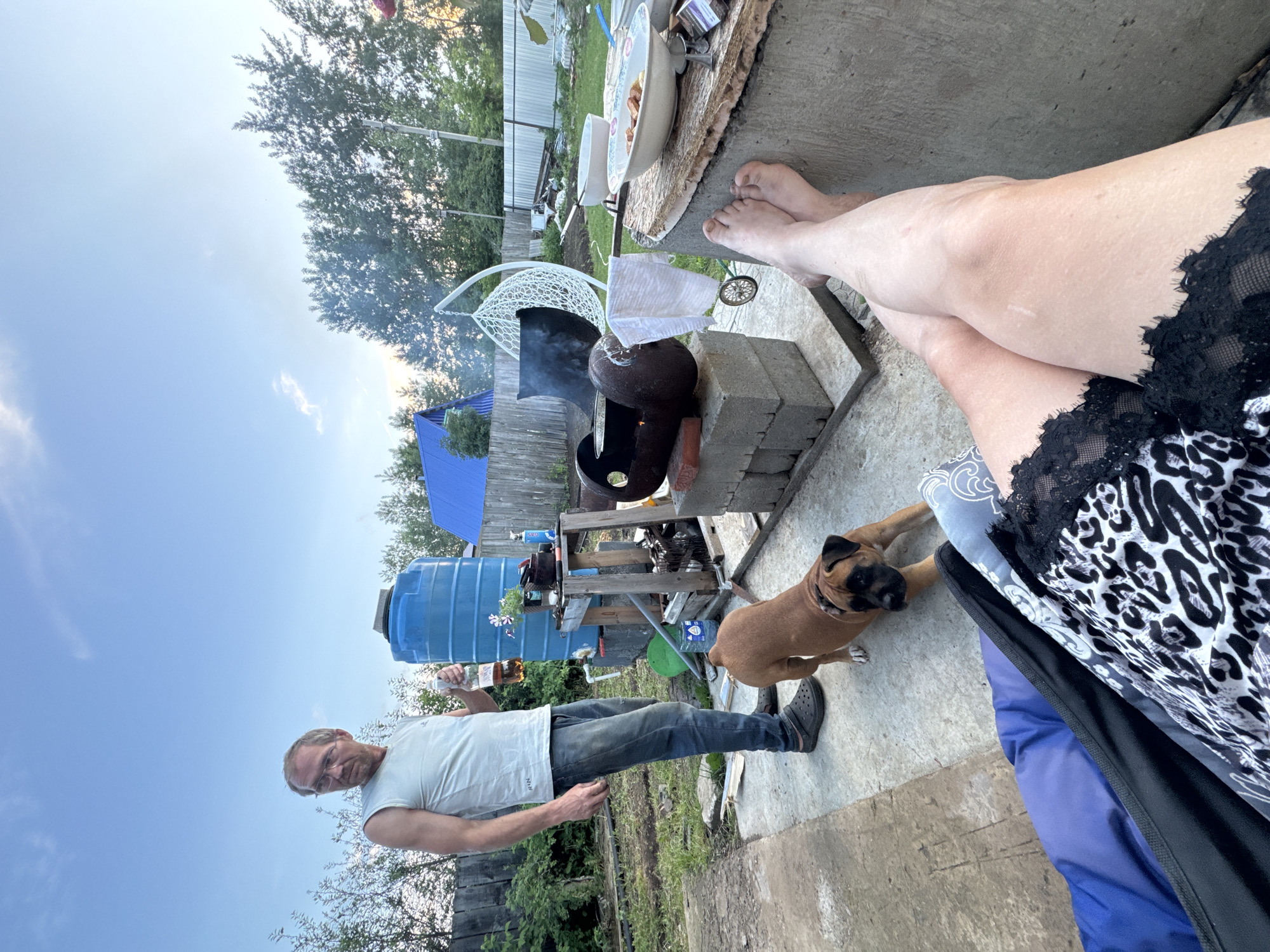 An outdoor scene shows a man in a white tank top and jeans holding a bottle of amber liquid, standing near a large blue water tank. A brown dog is in the foreground, looking towards the camera. Behind the man, a makeshift grill is set up with food cooking. A white hanging chair is visible in the background, along with trees and a fence. The ground appears to be concrete and dirt, suggesting a backyard or rural setting. The sky is blue with some clouds, and the overall lighting indicates it is daytime. The location is identified as Chaikovsky, Russia.