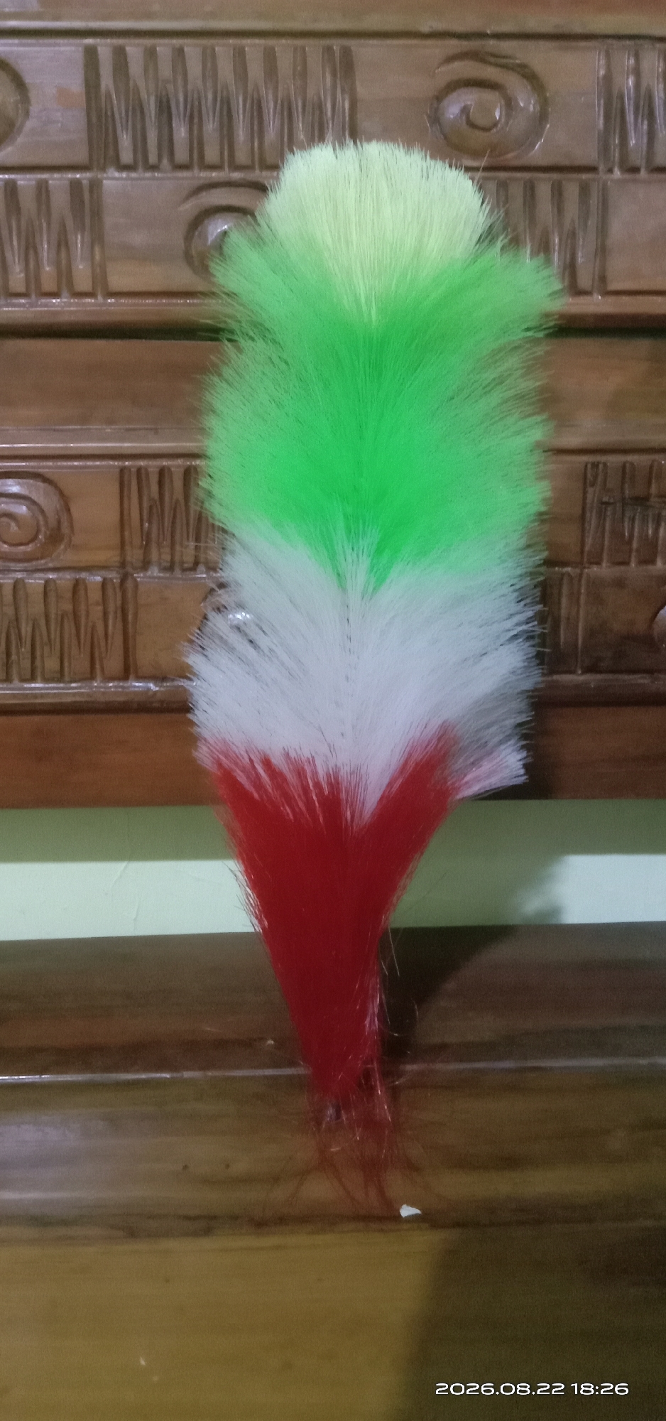 A fluffy, multi-colored duster, predominantly green, white, and red, stands upright against a wooden surface with an ornate, carved pattern. The duster's top section is light green, transitioning to white, and then to red at its base. The background is a textured, dark brown wood, possibly a piece of furniture, with horizontal plank lines and decorative carvings, including circular and rectangular motifs. Below the wooden surface, a pale green wall is visible. The overall scene is indoors and appears to be a still-life composition. The image was taken in Pakong, Indonesia.