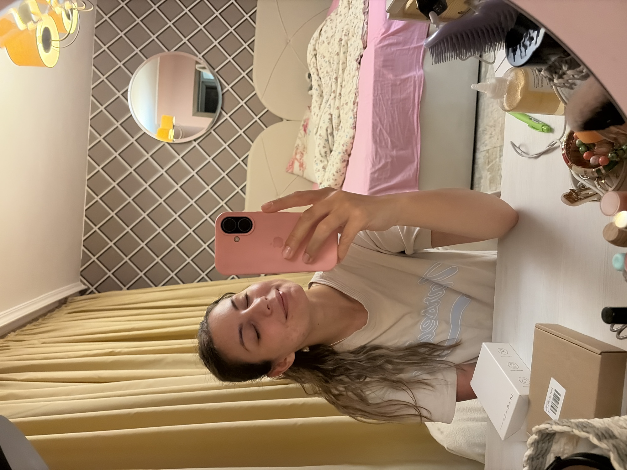 A woman is taking a selfie in a bedroom. She is holding a pink phone in her right hand and has her eyes closed and a slight smile on her face. She is wearing a light-colored t-shirt. In the background, there is a bed with a pink bedsheet and a floral comforter, and a patterned wallpaper. To the left of the woman, there are yellow curtains. On the table in front of her, there are some boxes and a basket with cosmetic items. The image appears to have been taken during the day, as suggested by the lighting. The location is identified as the city of Burgas, Bulgaria.