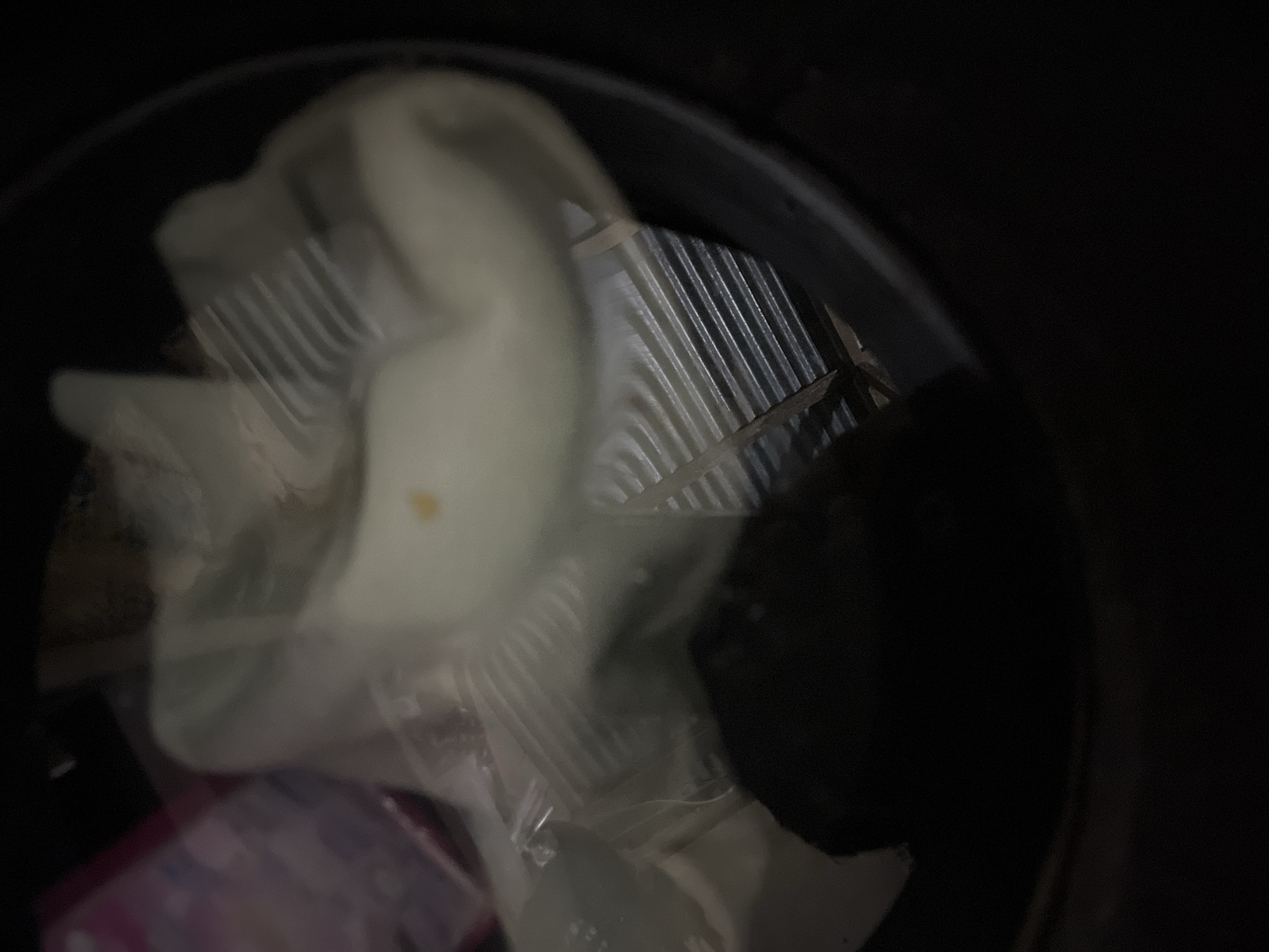 The image shows a close-up, top-down view of a dark, round container, likely a trash bin, filled with various items. A white, opaque object, possibly plastic or fabric, dominates the center, partially obscuring other contents. A few parallel lines, resembling a grate or vent, are visible beneath and within the white material. To the left, a portion of a pink and purple patterned object, possibly packaging, is visible. A small, brown speck or stain is present on the white object. The overall scene is dimly lit, with the focus on the contents of the container. There is no discernible context or detail to place this image within Benuaraja, Indonesia.