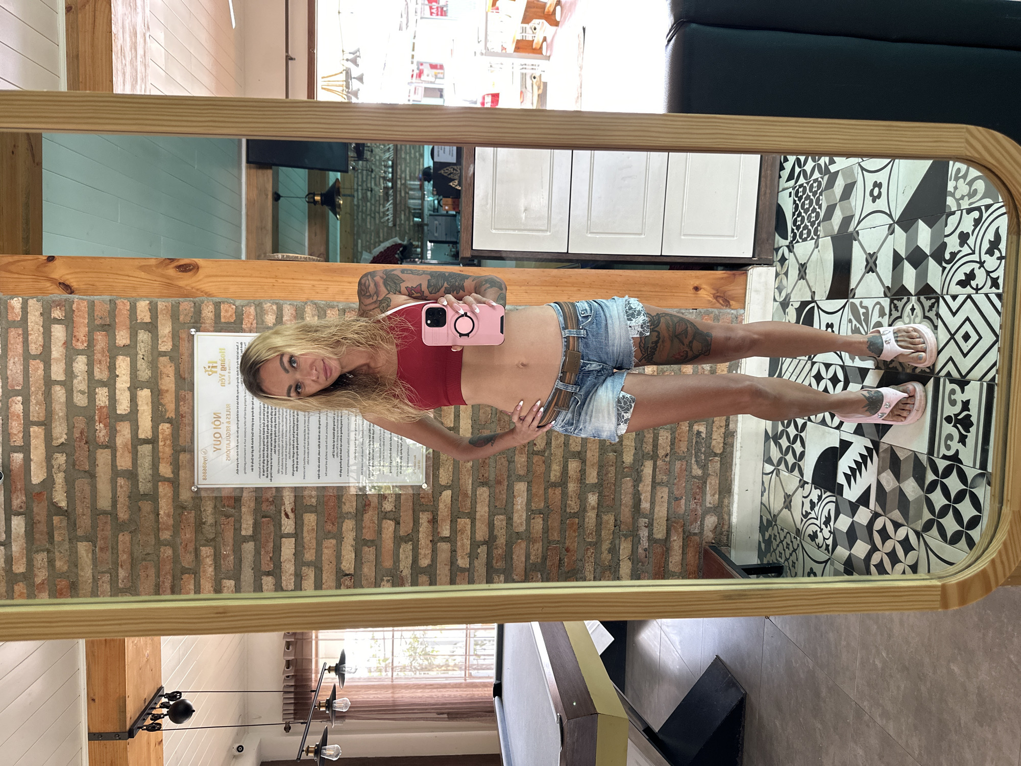 A woman, with long blonde hair and tattoos on her arms and legs, poses for a mirror selfie in what appears to be a cafe or restaurant in Da Lat, Vietnam. She is wearing a red crop top, denim shorts, and pink flip-flops. She is holding a pink smartphone in her right hand. In the background, a brick wall with a sign in Vietnamese is visible, as well as a pool table and various decorative elements. The floor is covered in a black and white patterned tile.