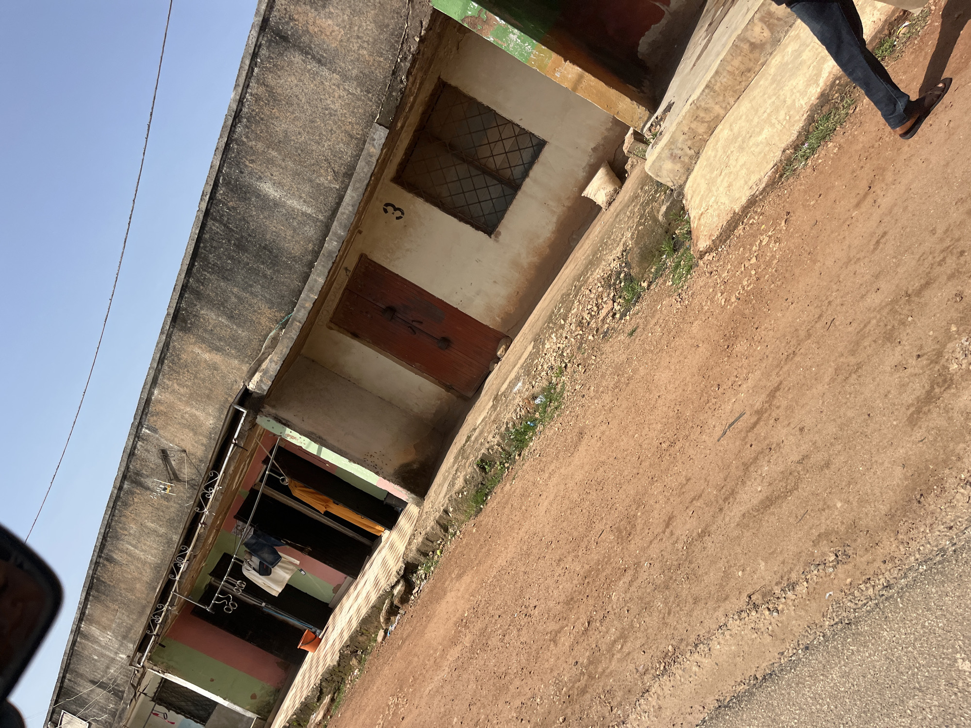 The image depicts a street scene in Gura Top, Nigeria. A narrow, unpaved dirt road runs diagonally across the foreground, transitioning to a paved concrete sidewalk in front of a low-rise building. The building has a weathered, concrete exterior and is painted in sections with stripes of pink, green, and white. Several doors and a barred window are visible. Laundry hangs on a line in front of some of the doorways, and an orange bucket sits on the sidewalk. The sky is clear and blue. In the lower right corner, a person's legs and feet are visible, wearing dark jeans and brown sandals, as they walk away from the camera along the sidewalk.