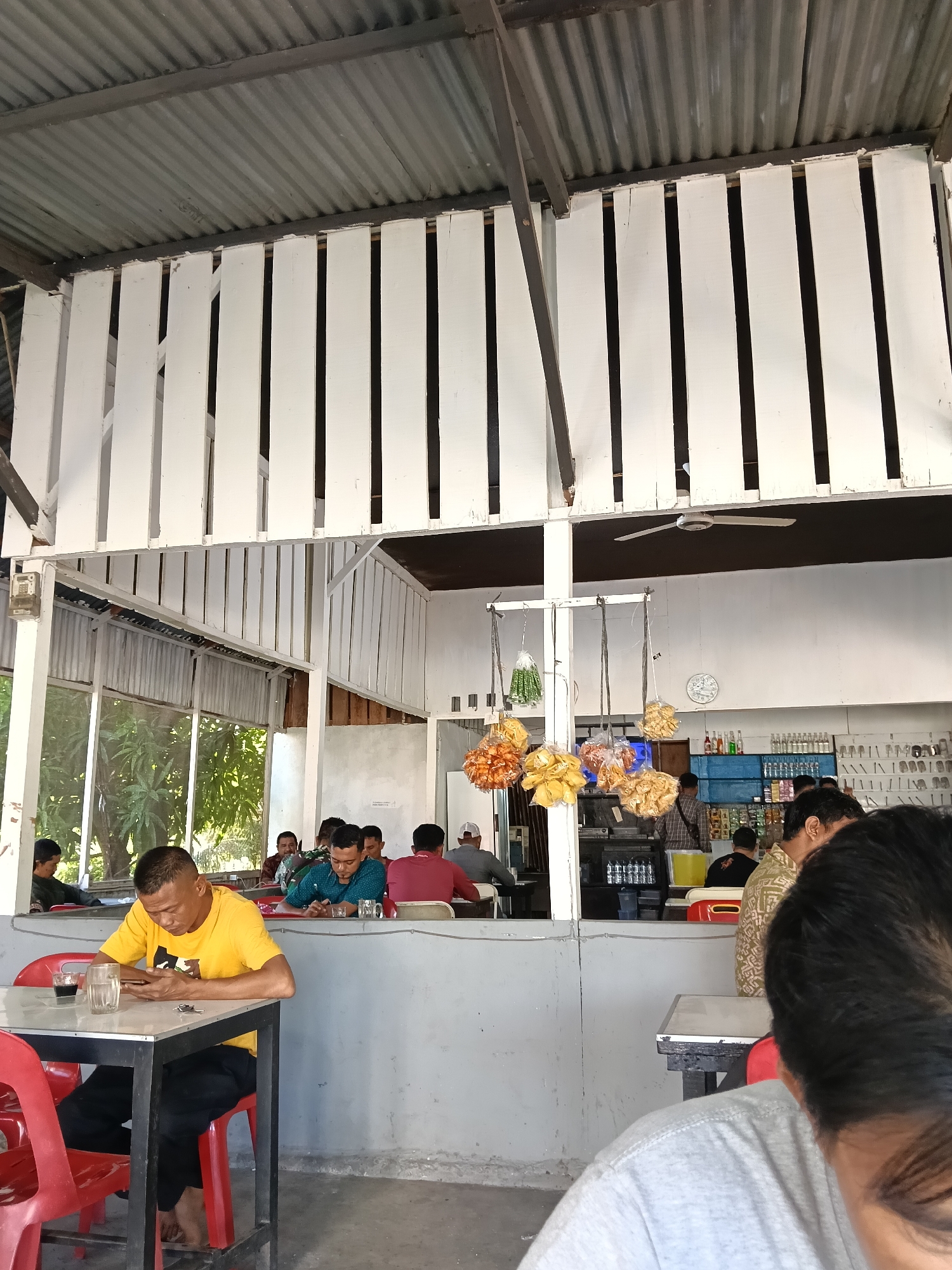 The image shows an indoor seating area, possibly a restaurant or warung, in Banda Aceh, Indonesia. The ceiling is constructed of corrugated metal and white vertical wooden planks. The walls are painted a light gray. In the foreground, a man wearing a yellow t-shirt and black pants is sitting at a white table with black legs, looking at his phone. He has a glass of dark liquid and a small object on the table. Red chairs are visible at the table.
Further back, several other people are seated at tables. To the left, through an open window or doorway, trees and foliage are visible, suggesting an outdoor area or a proximity to nature. The background includes a counter area with shelves stocked with various bottles and items, and a clock on the wall. Several bags of snacks are hanging from a structure in the middle of the seating area. The overall scene depicts a casual, communal gathering space.