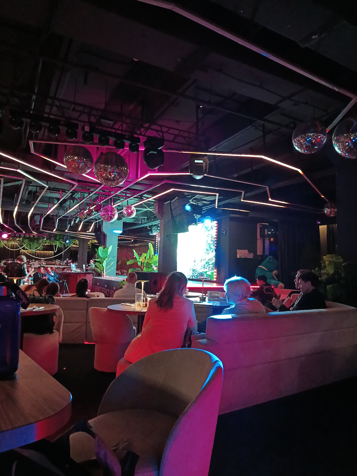 The image captures a scene inside what appears to be a lounge or nightclub, possibly in Moscow, Russia, as indicated by prior knowledge. The interior is dimly lit, illuminated by ambient lighting from pink neon light fixtures in the ceiling, disco balls, and the glow of a large video screen on a stage. Patrons are seated in upholstered furniture, including couches and individual chairs, grouped around tables. People are engaged in conversations. In the background, a bar area is visible. The atmosphere is social and oriented toward entertainment.