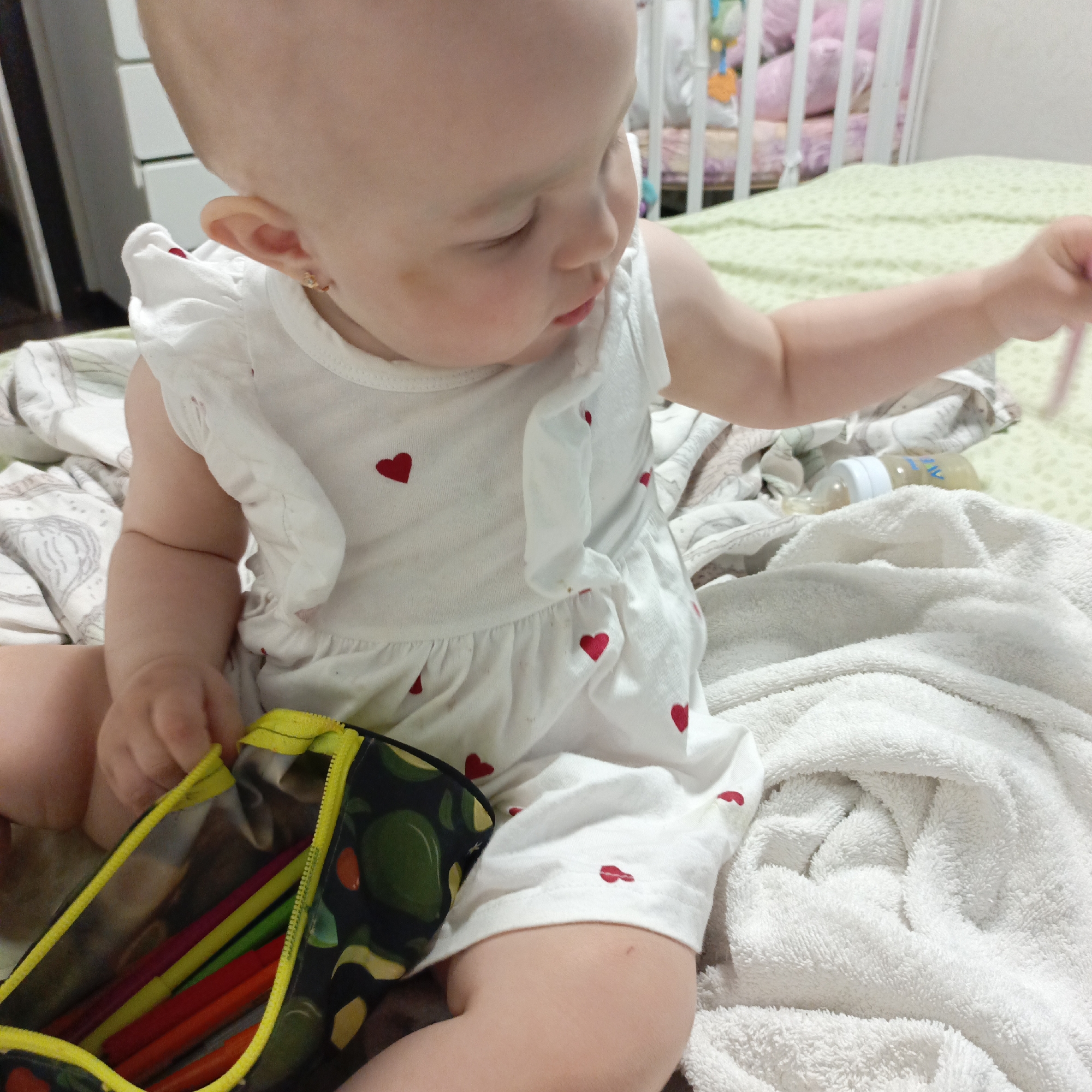 A baby is sitting on a bed. She is wearing a white dress with red hearts. She has earrings. In her left hand, she is holding a pencil case with colored pencils. Next to her is a baby bottle. The bed has a green sheet and a white blanket. Behind the baby is a white crib.