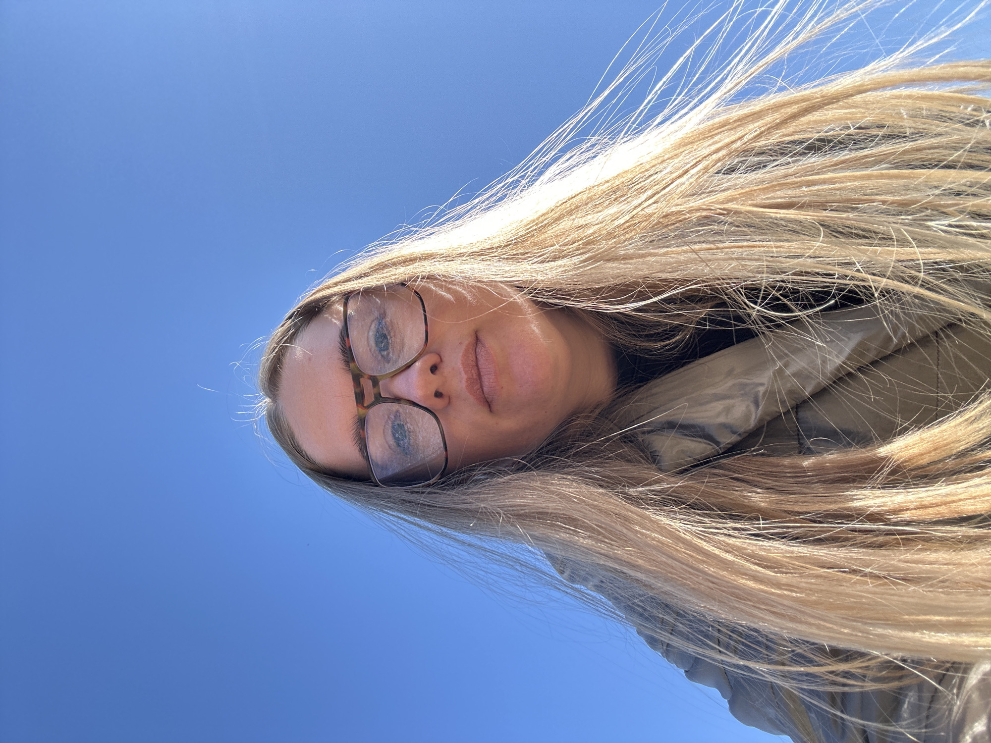 An angled upward shot captures a fair-skinned woman with long, blonde hair and glasses against a bright blue sky. Her hair is windblown, obscuring some of her face and part of her clothing, which appears to be a jacket or coat. The location cannot be determined to be in Уральское, Russia based on the information available.