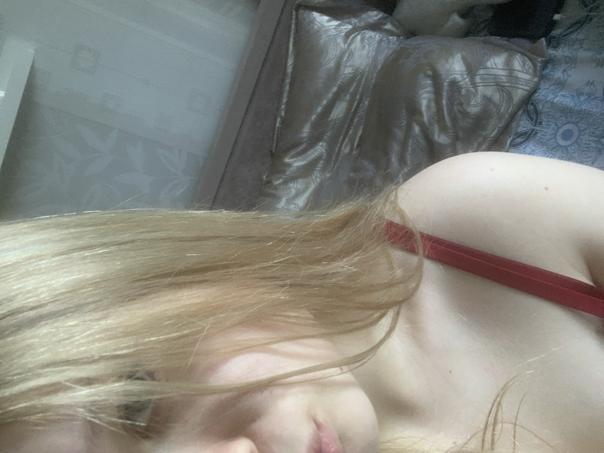 A close-up, skewed-angle image shows a person with blonde hair lying down, partially visible face with lips, fair skin, and a red bra strap. The background includes bedding with a shiny, patterned pillow and a wall with a subtle leaf pattern. No location details of Saint Petersburg, Russia are discernible from the image.