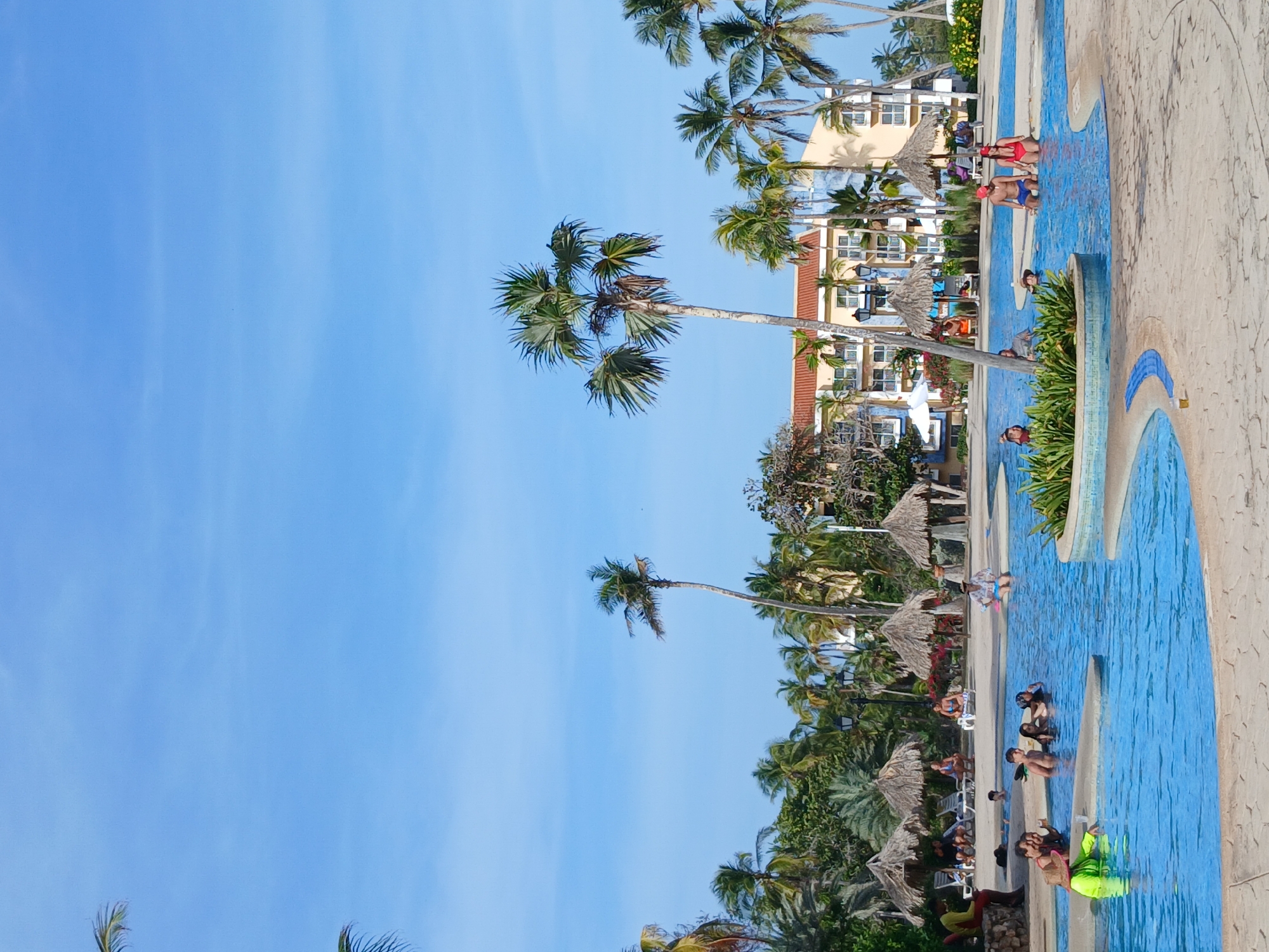 A view of a resort in Playa El Agua, Venezuela shows a swimming pool with several people in the water. Some are sitting on the side of the pool. A few are standing near the pool, some wearing red and blue bathing suits. Several palm trees stand around the pool. Thatched umbrellas are near the pool and lawn chairs. A multi-story hotel is set back from the pool. The sky is blue with a few clouds.