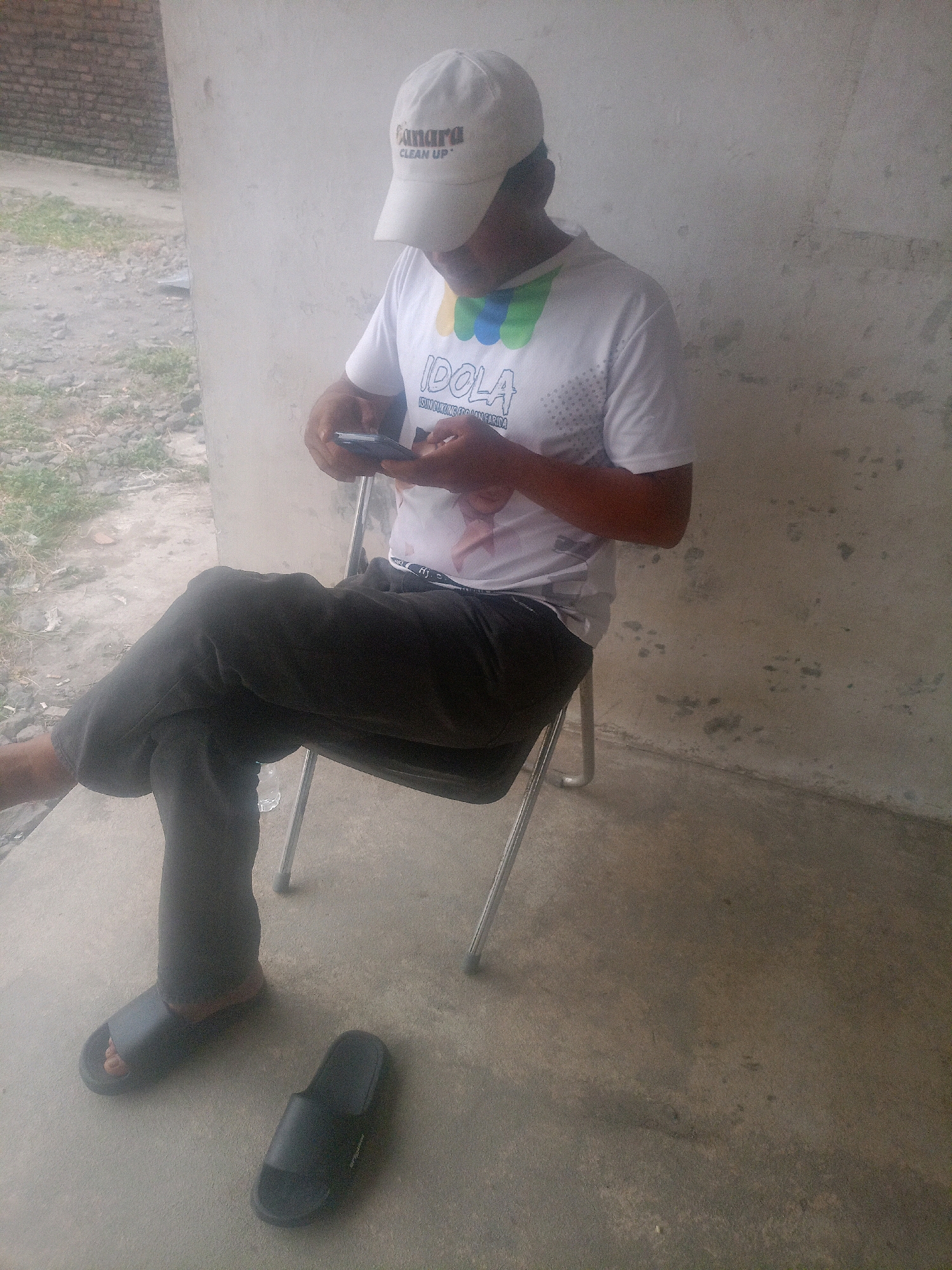 A person is sitting on a metal chair with a white baseball cap and a white t-shirt, holding a mobile phone. The person is wearing dark pants and black sandals. Another black sandal is placed on the ground next to the seated person. The background consists of a textured wall and a concrete floor. The image appears to be taken outdoors, possibly in Cirebon, Indonesia, given the context.