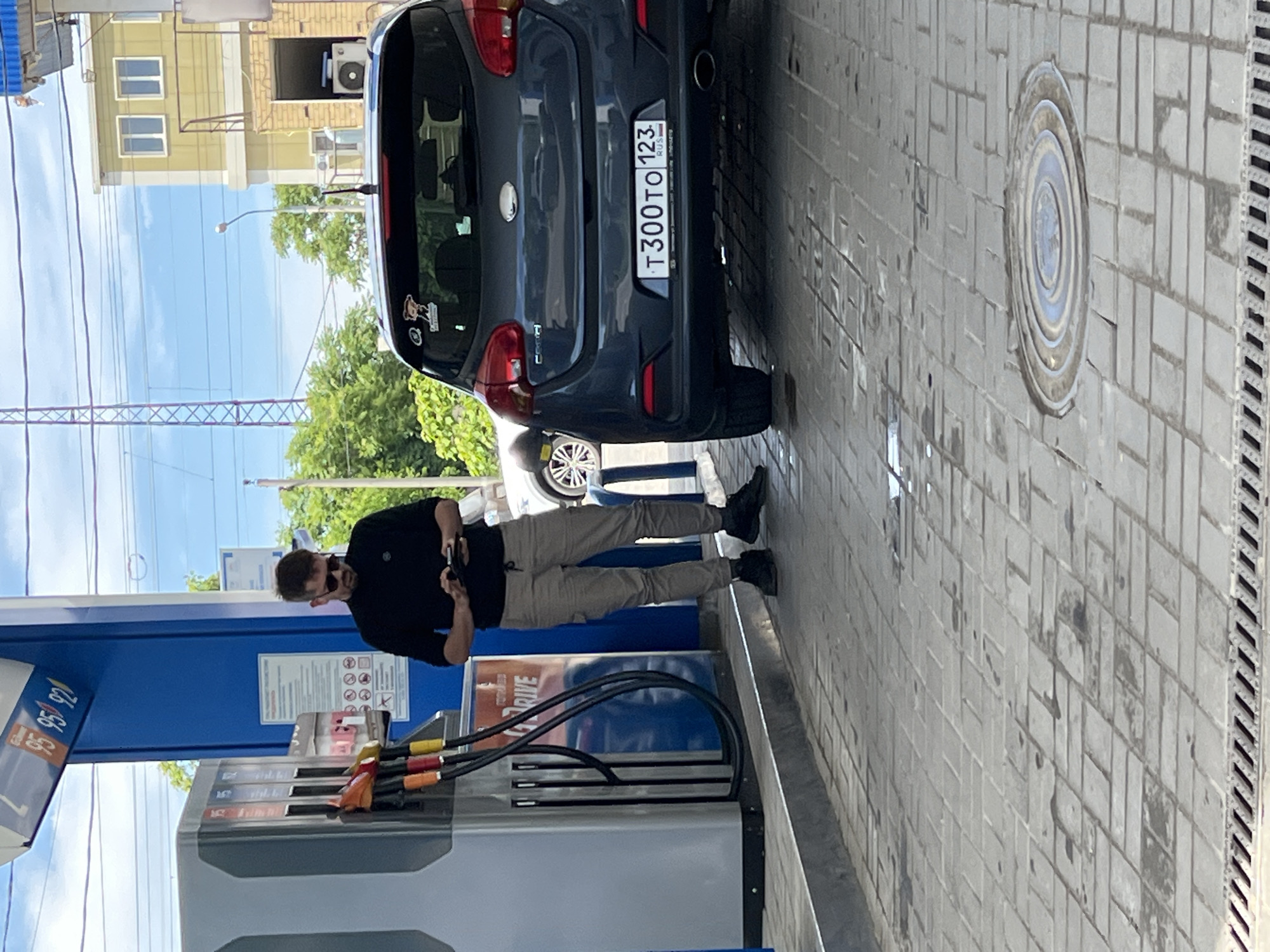 This image depicts a daytime scene at a gas station, likely in Krasnodar, Russia, given the license plate details. A man, dressed in a black long-sleeved shirt, light beige trousers, and black shoes, stands near a blue and grey fuel pump. He is wearing sunglasses and appears to be occupied with his phone, holding it with both hands, while standing beside the fuel dispenser.
A dark grey hatchback car is parked at the pump with its rear facing the viewer. The car's license plate, "T300TO 123 RUS," clearly indicates its registration in Russia, with "123" being the code for Krasnodar Krai. A couple of small stickers are visible on the car's rear window, one of which partially reads "СЧАСТЬЕ В..." (Happiness in...).
The fuel pump displays "95" and "92," indicating different octane ratings available. Various colored fuel nozzles are visible, hanging from the pump. The ground around the pump is paved with grey interlocking tiles, and a circular manhole cover is present to the right. In the background, under a bright, clear sky, power lines stretch across the frame, and lush green trees are visible. A light yellow building with white-framed windows and an external air conditioning unit can be seen in the distance. The overall weather appears sunny. A sign on the fuel pump structure shows common gas station warnings, including symbols for no smoking and no phone use.