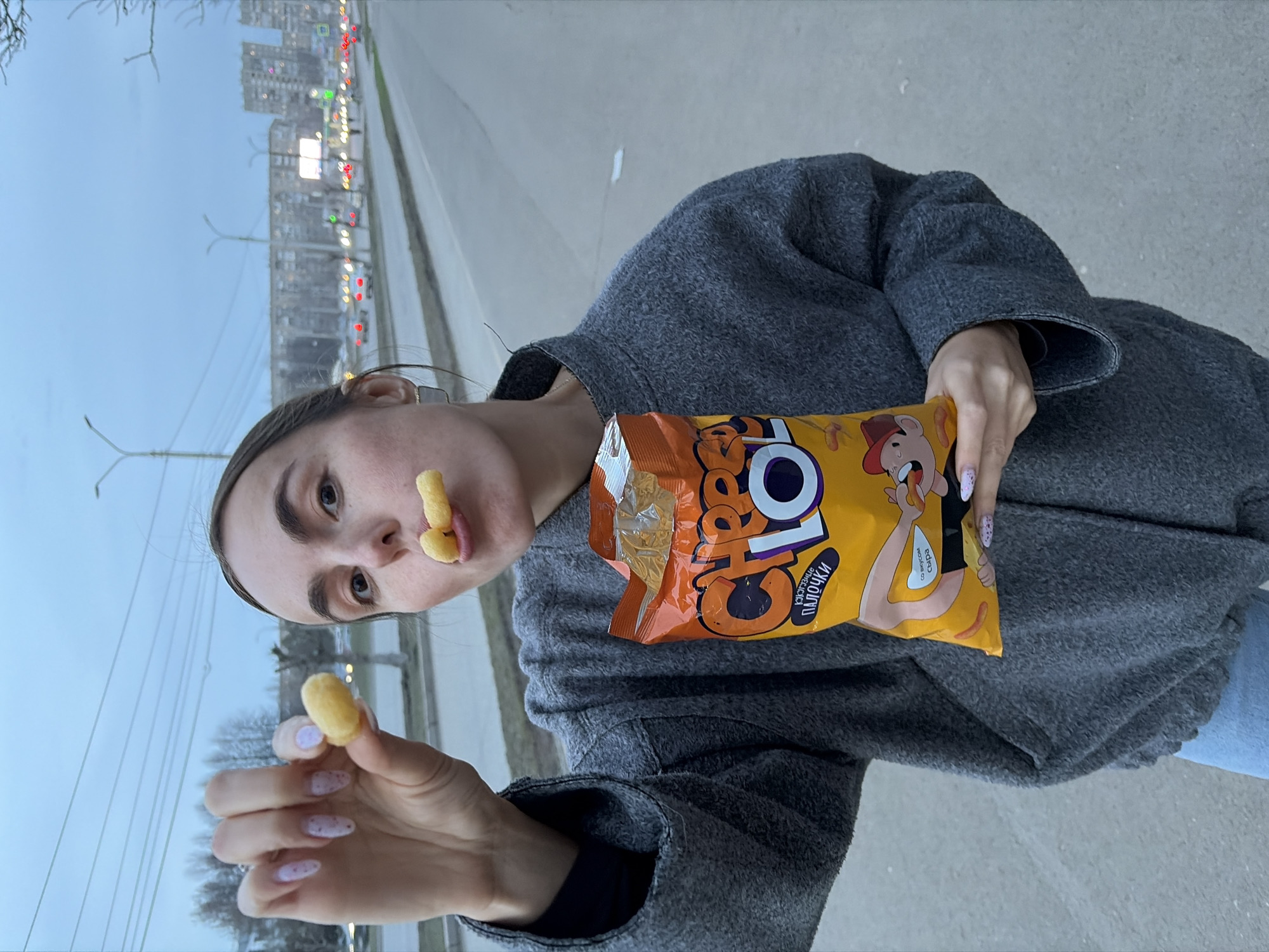 A woman stands outside in Izhevsk, Russia, holding a bag of Cheetos brand cheese puffs. One cheese puff is held between her fingers, and another is stuck on her upper lip, partially obscuring her mouth. She wears a gray wool coat. Behind her is a city street with buildings in the distance, street lights, and car lights. The time of day appears to be dusk.