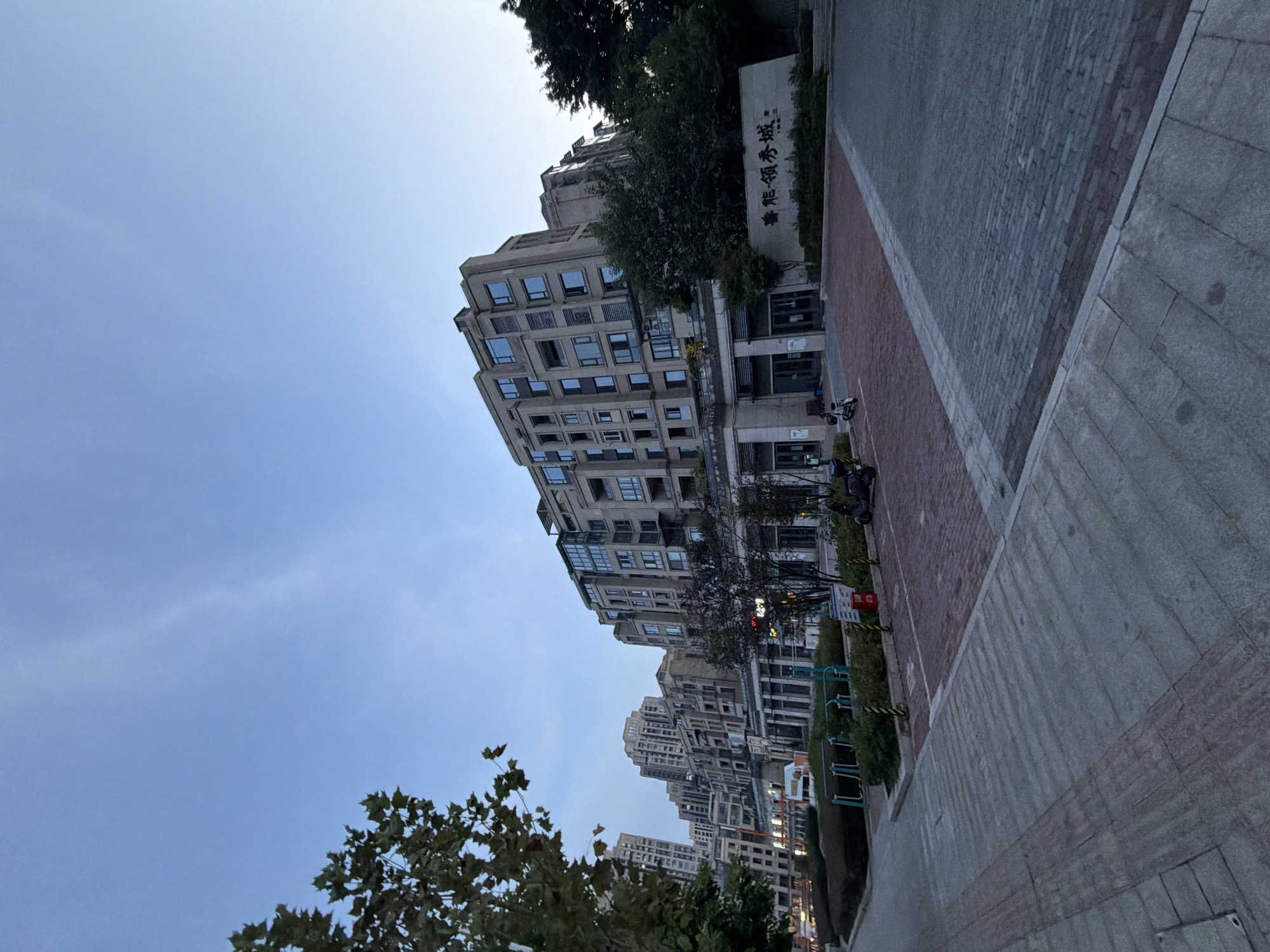 The image depicts a wide-angle view of a modern residential complex in Jiangjin, China, during the late afternoon or early evening. The sky is a clear, pale blue with faint wisps of clouds. In the foreground, a paved sidewalk runs diagonally from the lower left to the mid-right of the frame. This sidewalk is constructed with large, rectangular grey stone slabs. Adjacent to the sidewalk, to the right, is a driveway or parking area paved with red brick.
The main subject of the image is a multi-story apartment building with a light-colored facade. The building features numerous windows, many of which are recessed, and balconies. The architecture appears contemporary, with clean lines and geometric shapes. In front of the building, there is a low-rise section with a glass-enclosed facade and columns, suggesting commercial or communal spaces on the ground floor.
To the right of the entrance to the building, a large stone monument or sign displays Chinese characters, likely indicating the name of the residential complex. Lush green trees and shrubs are visible around the building and along the perimeter of the complex, with some taller trees framing the left side of the image.
Several objects are scattered around the entrance area. Two electric scooters are parked near the building's entrance. There are also some bicycle racks and a red waste bin. In the background, other buildings of varying heights can be seen, suggesting a densely populated urban environment. The lighting suggests it is dusk, as the light is soft and there are no harsh shadows, and some windows in the buildings appear to be illuminated. The overall scene conveys a sense of urban residential living in a well-maintained development.