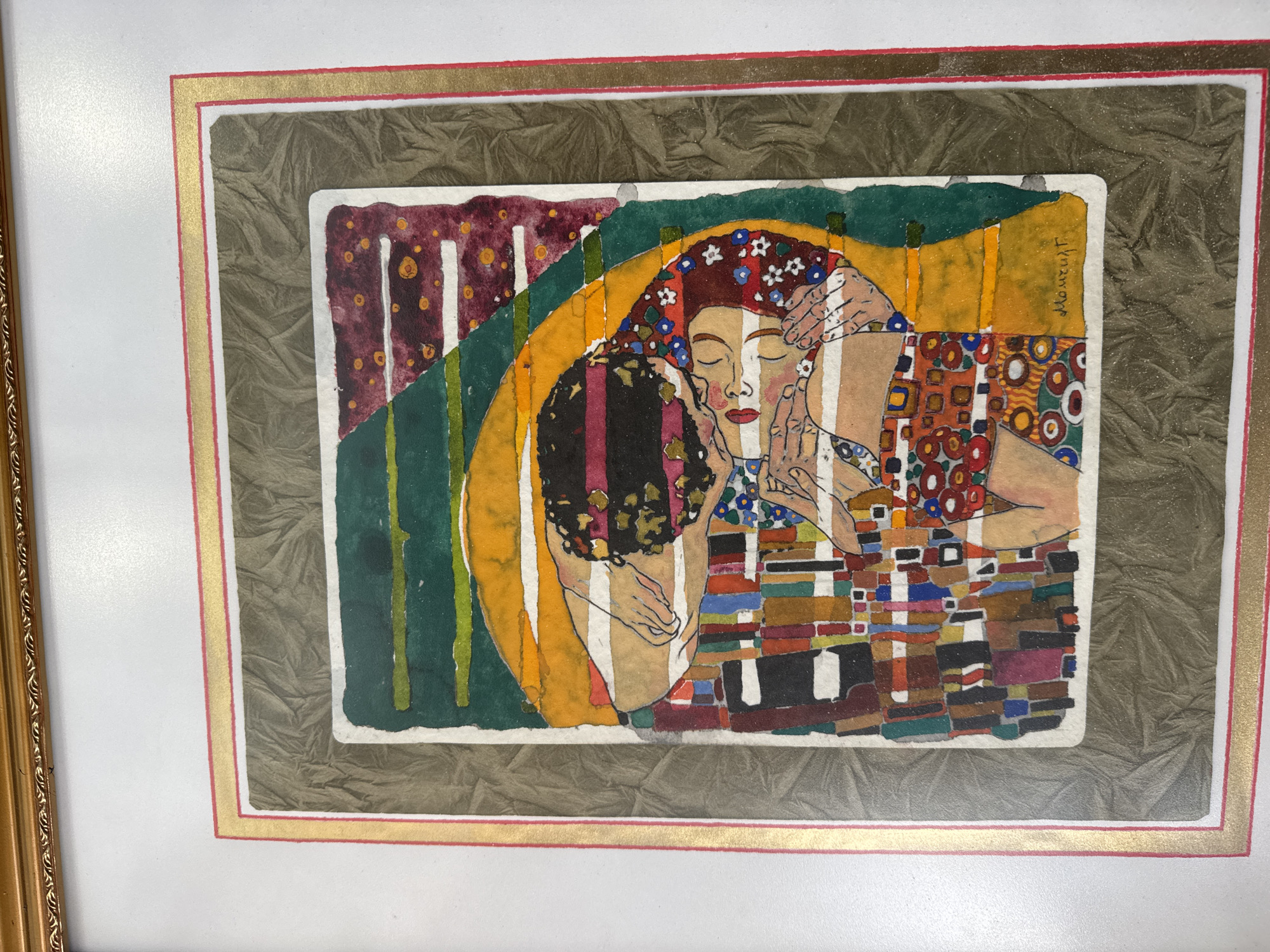 The image is a framed watercolor painting. The painting depicts two figures in an embrace, reminiscent of Gustav Klimt's "The Kiss". The figures are partially obscured by geometric patterns in various colors and shapes, similar to Klimt's style. The woman has her eyes closed and a floral head covering. One figure is dark-haired. The background features vertical stripes in colors like maroon, green, and yellow. The painting is signed in the upper right corner. The artwork is framed with a white border, followed by a brown, textured border, and an outer gold frame. The image does not contain any information about the city of Sofia, Bulgaria, nor does it offer any information about the location in Sofia, Bulgaria.