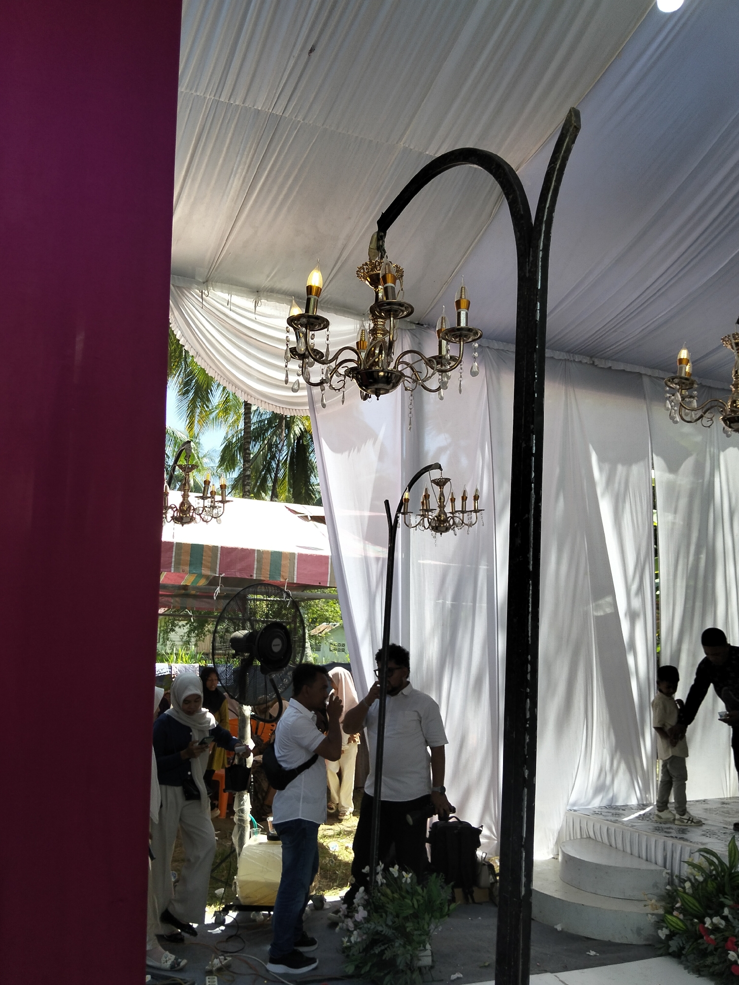 The image depicts an outdoor event setup under a white tent canopy. The scene is likely in Pulonga, Indonesia, as suggested by the presence of palm trees visible through openings in the tent. In the foreground, to the left, a deep magenta drape forms a vertical boundary.
The tent's ceiling is draped white fabric, and several ornate brass chandeliers hang from it. One prominent chandelier is attached to a curved black metal pole.
Several people are present. On the left side, a woman wearing a white hijab and light-colored pants is looking at her phone. To her right, a man in a white short-sleeved shirt and blue jeans stands facing away from the camera, holding a camera bag. Further to the right, a young boy in a light-colored shirt and pants stands on a raised circular platform, interacting with a man in a dark shirt who is also facing away from the camera.
In the middle ground, an industrial fan is visible. Other people, partially obscured, are seen in the background. There are floral arrangements in the lower right corner. The ground appears to be covered with a patterned mat or cloth. The overall impression is of a partially decorated event space, possibly for a wedding or celebration.