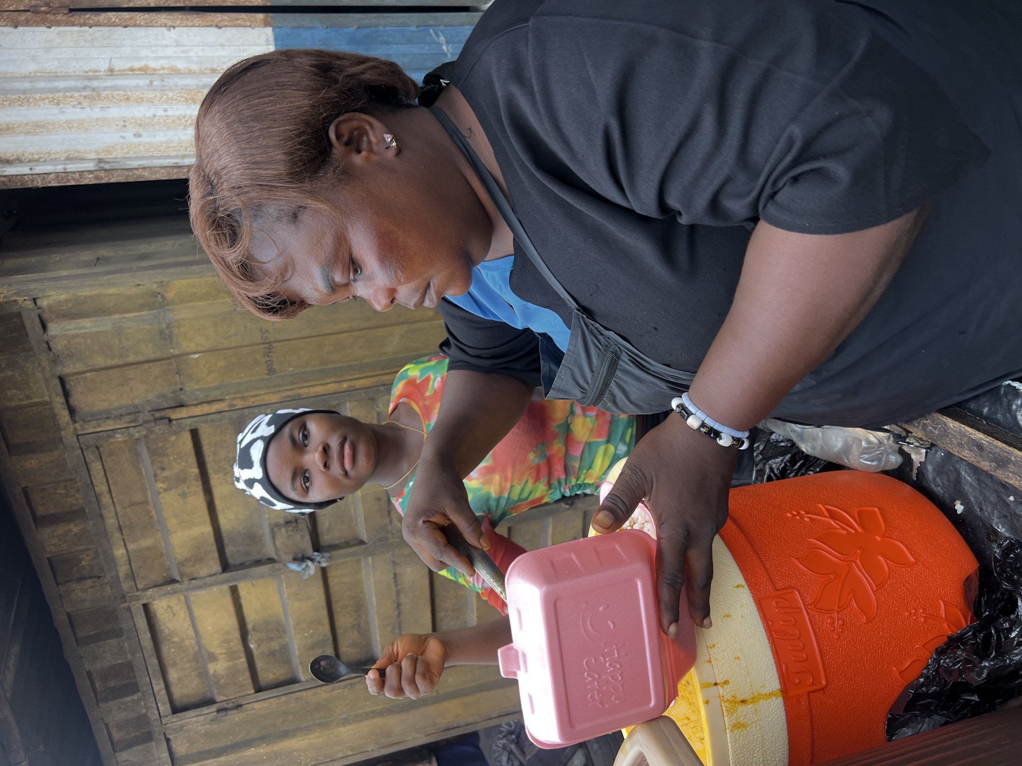 The image shows two people at a street food stall in an unknown location. An older woman with brown hair and a black shirt is using a pink container to scoop something from a large orange and cream cooler labeled "Tulip". A younger woman with a black and white headscarf and a floral shirt stands nearby, holding a spoon. The background consists of corrugated metal and wooden panels.
