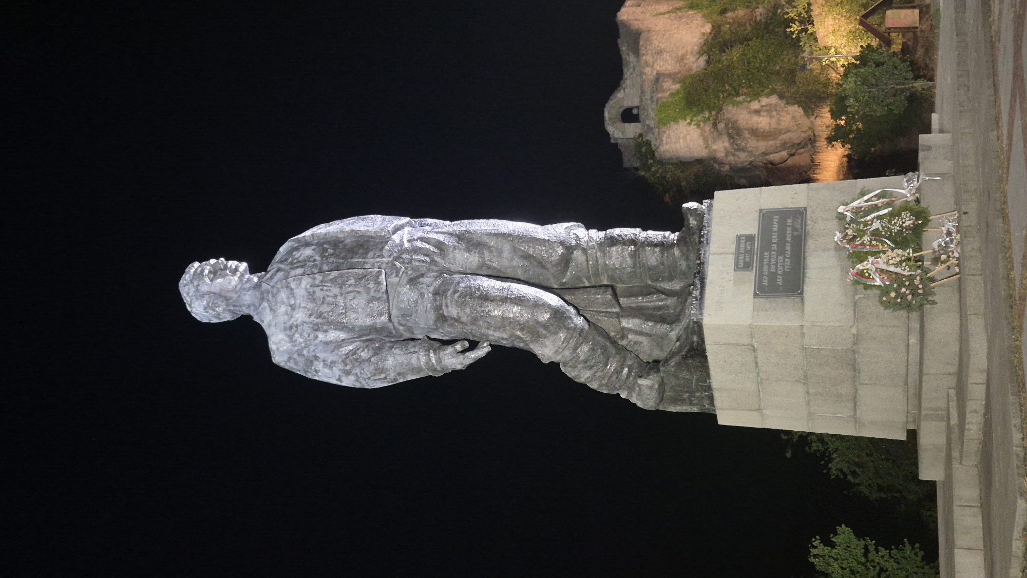 A full-body, weathered statue of a man, likely a historical figure given the surrounding inscription and monument, stands illuminated against a dark night sky. The statue is made of a dark, possibly bronze or stone material, with a slightly metallic sheen reflecting the artificial light. The figure is depicted in a dynamic pose, with one leg forward and bent, and the other extended behind. He is wearing what appears to be a jacket or uniform with visible folds and creases, and his pants are tucked into boots. The statue is mounted on a large, multi-tiered concrete pedestal.
On the front of the pedestal, there is a metal plaque with an inscription in Cyrillic script, followed by a smaller inscription. Below the plaque, three memorial wreaths with red and white ribbons are placed on the steps of the pedestal. To the right of the statue and pedestal, a stone ruin with arched openings, partially covered in ivy, is visible on a rocky incline. Green foliage surrounds the base of the ruins and the monument. A small wooden structure, possibly a signpost or information kiosk, is also visible in the lower right portion of the image. The lighting suggests the statue and its immediate surroundings are the primary focus, with the background ruins faintly illuminated by spotlights.
This statue is located in the city of Lovech, Bulgaria, specifically on the Varosha hill. The inscription on the plaque likely refers to a prominent historical figure associated with the city or Bulgaria. The statue is a key landmark in Lovech, situated in a historically significant area of the city.