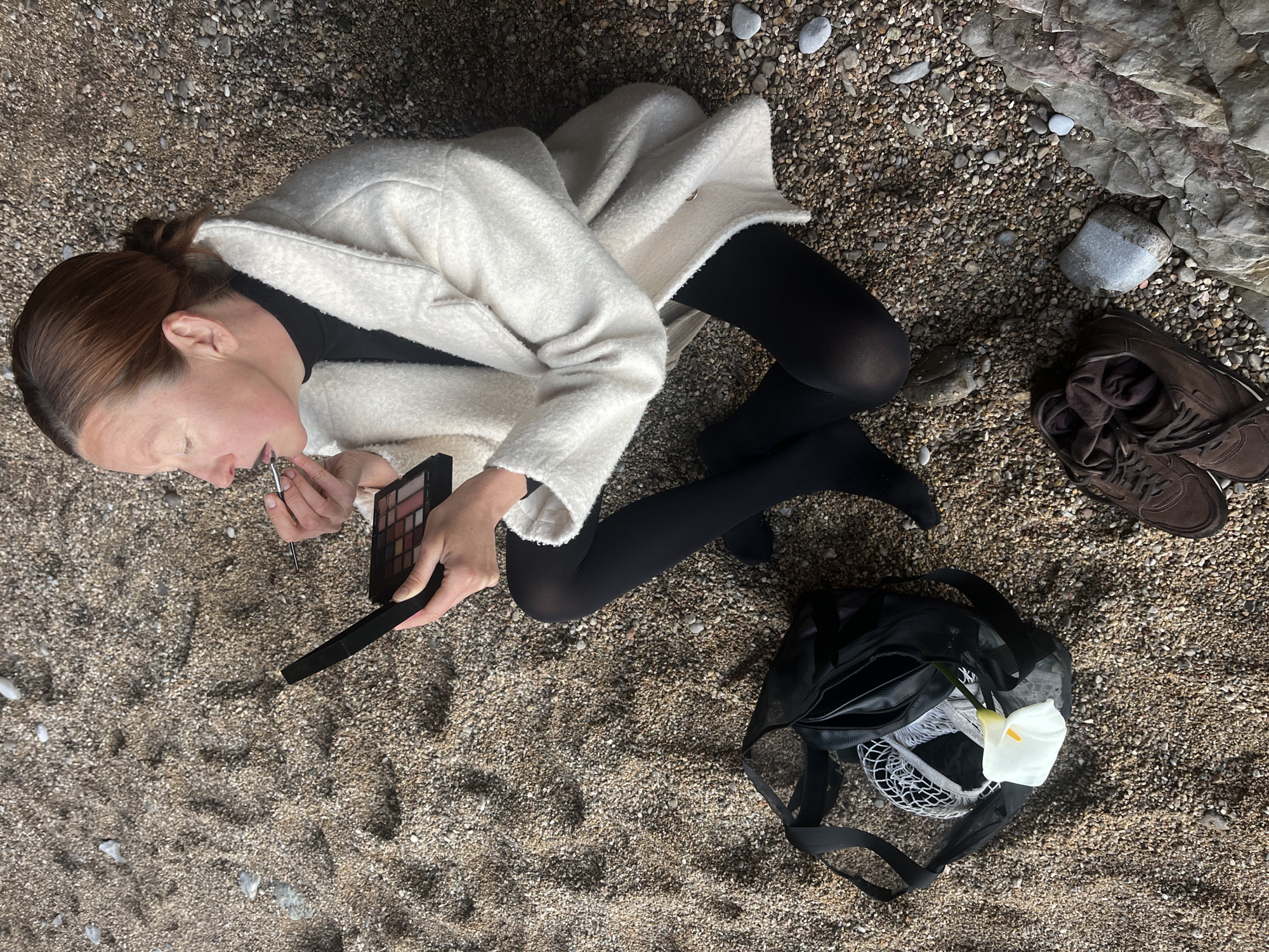 A woman sits on a beach in Budva, Montenegro, applying makeup. She wears black tights, a black top, and a white jacket. A makeup palette and brush are in her hands. Next to her is a black mesh bag containing a white calla lily and a white net bag. A pair of brown athletic shoes lies nearby, and pebbles and a rock formation are visible in the background. The ground is covered in sand and small pebbles.